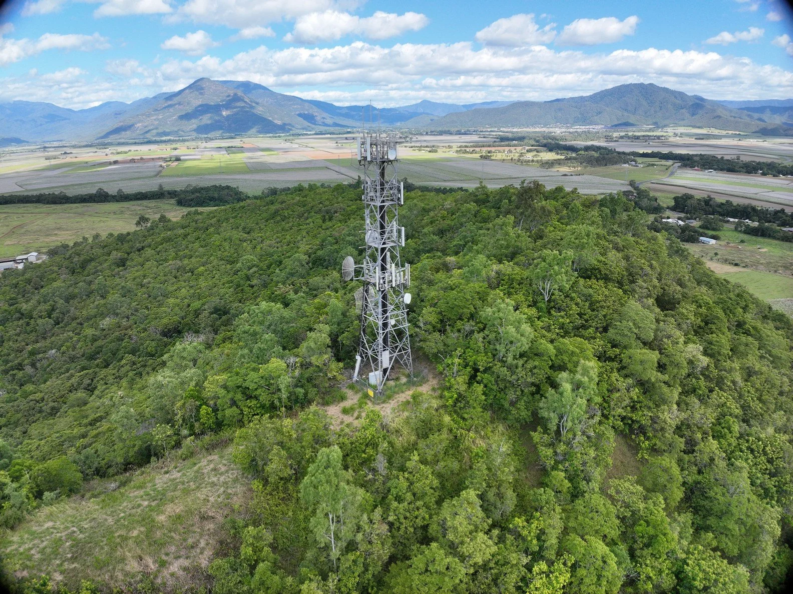 A metal radio tower positioned on a densely wooded hilltop with a backdrop of distant mountains and farmland.