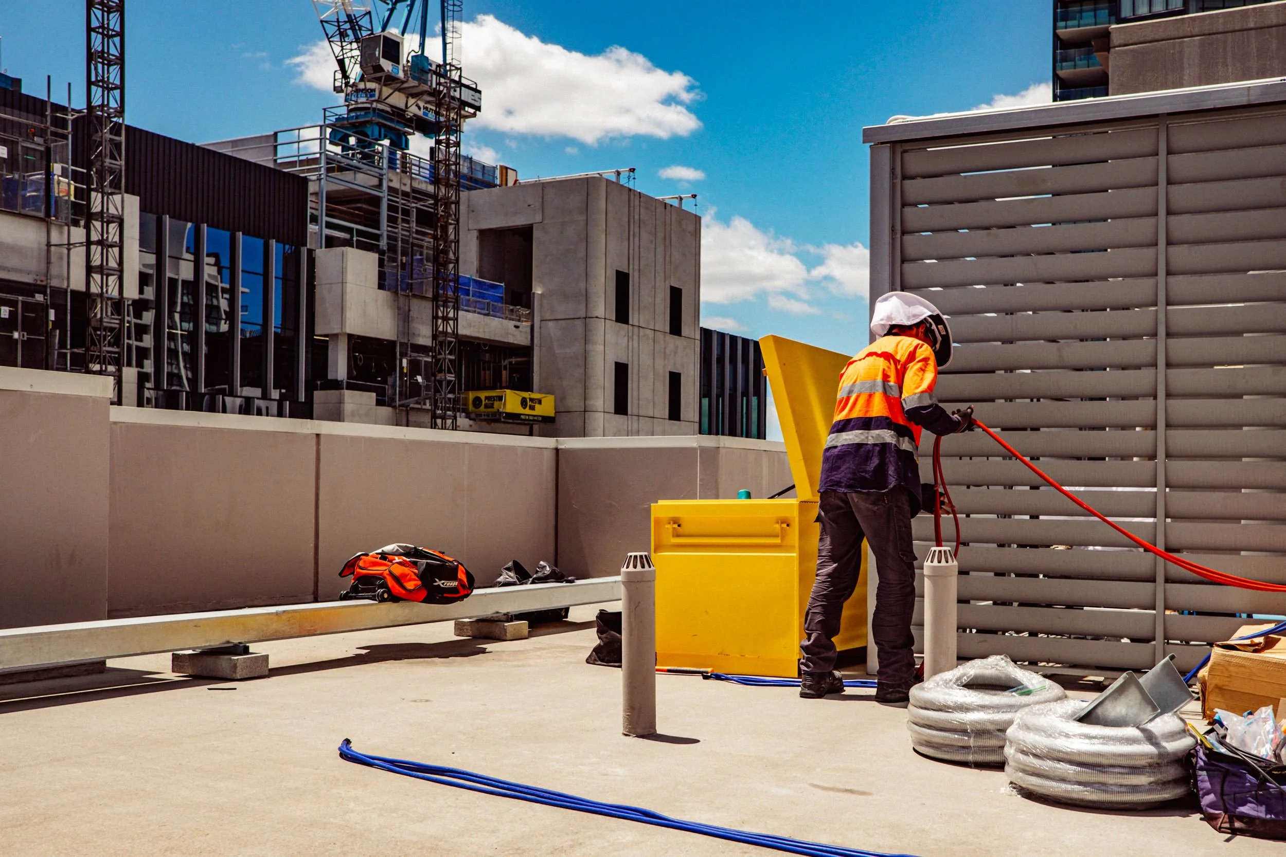 A construction worker in safety gear working with a yellow equipment box on a rooftop, with construction site in the background.