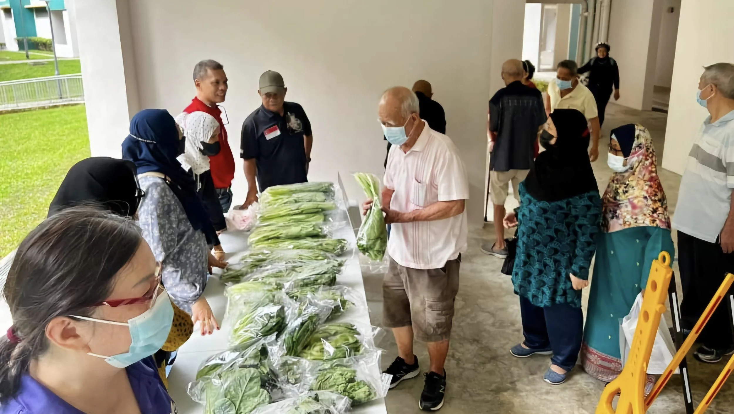 People wearing face masks standing in line at a vegetable stall with fresh green vegetables on a table.