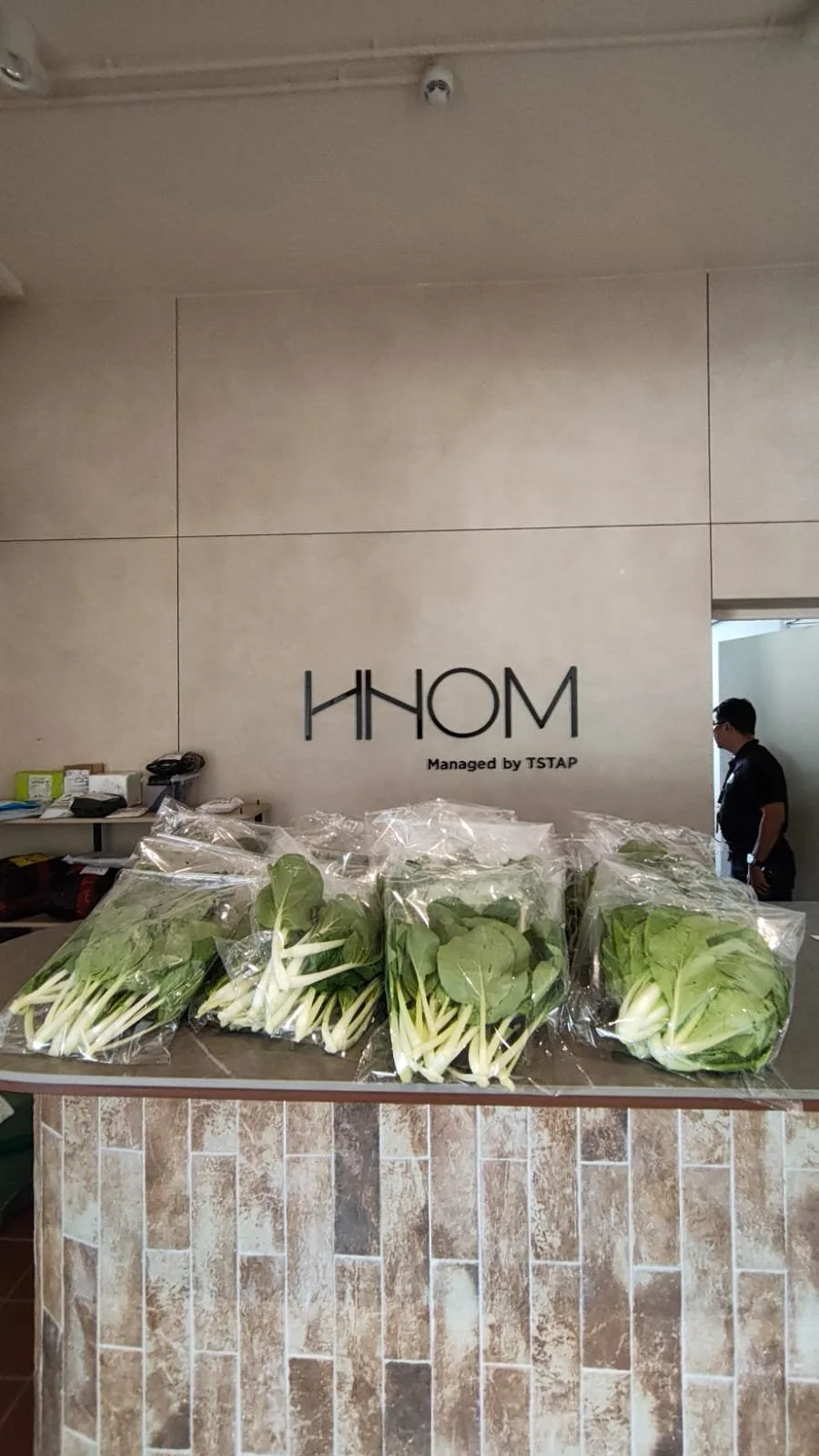Fresh leafy greens packaged in clear plastic bags placed on a counter with a tiled front at a hospitality or retail establishment, with the walls displaying the logo 'HHOM' and indicating it is managed by TSTAP.