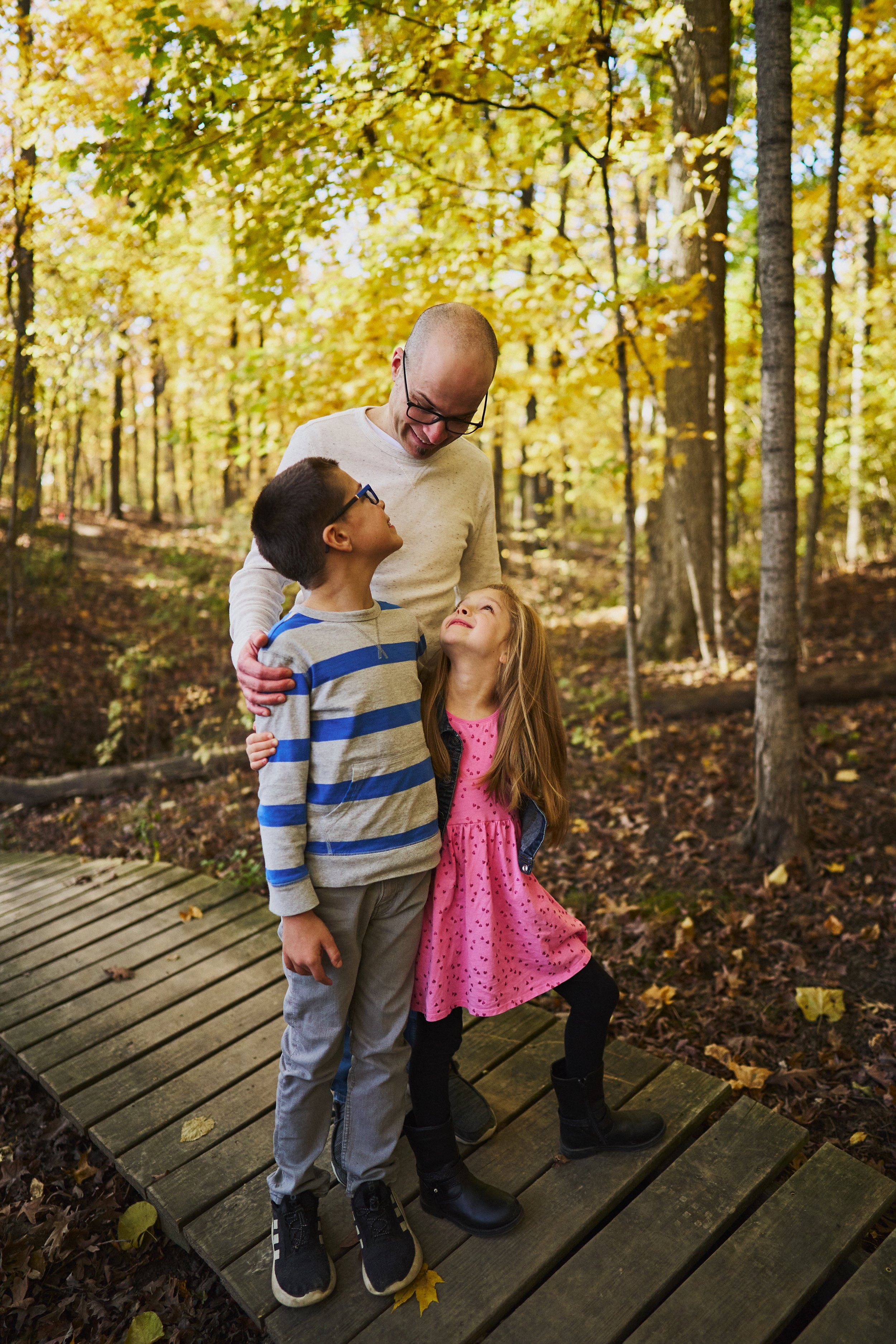 A man with glasses and two children, a boy and a girl, standing on a wooden path in a forest during autumn. The man is smiling and looking down at the children, who are looking up at him. The boy is wearing a striped sweater, and the girl is in a pin