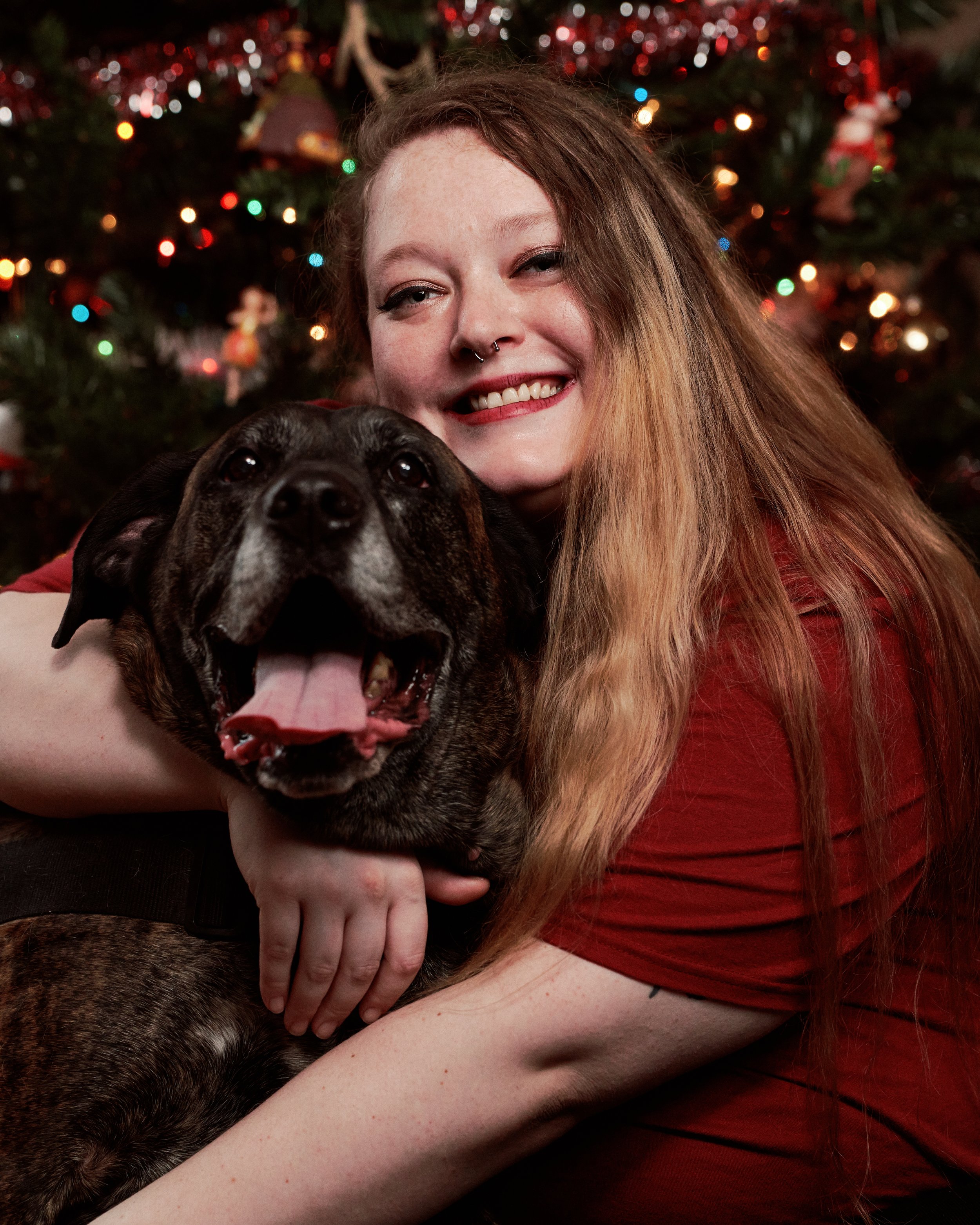 Woman smiling with long hair and nose ring, hugging a happy, panting dog in front of a decorated Christmas tree.
