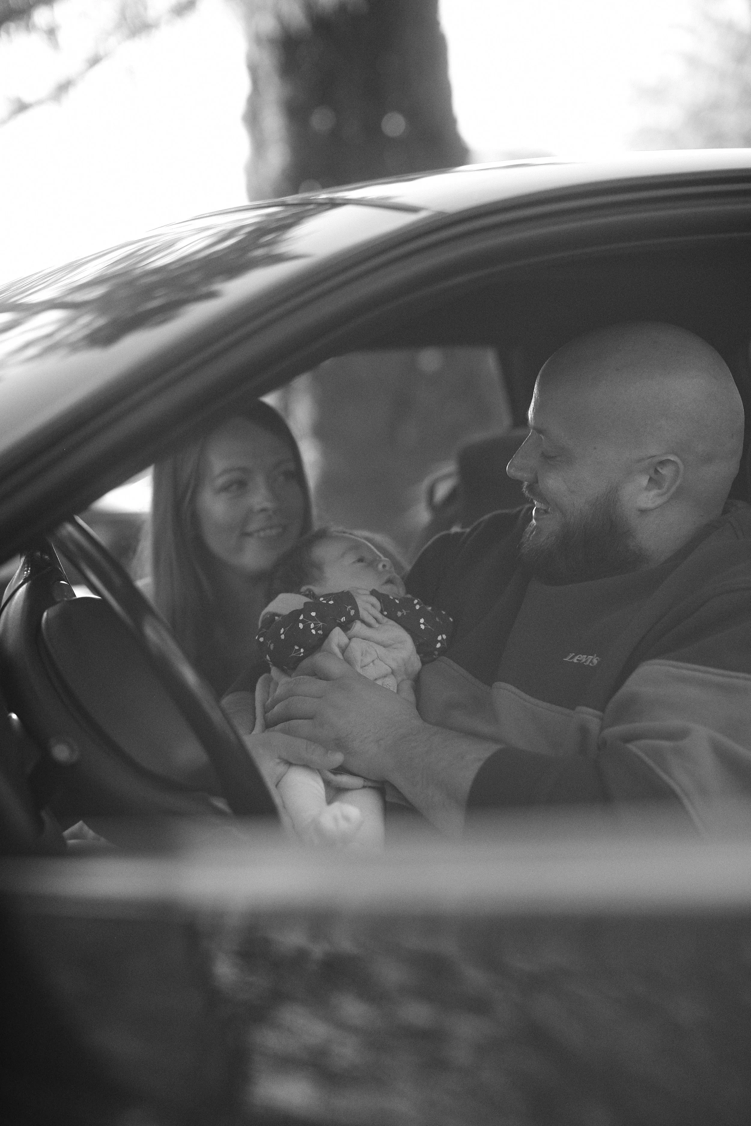 Black and white photo of a couple sitting in a car, with the man holding a baby and both smiling at the infant.