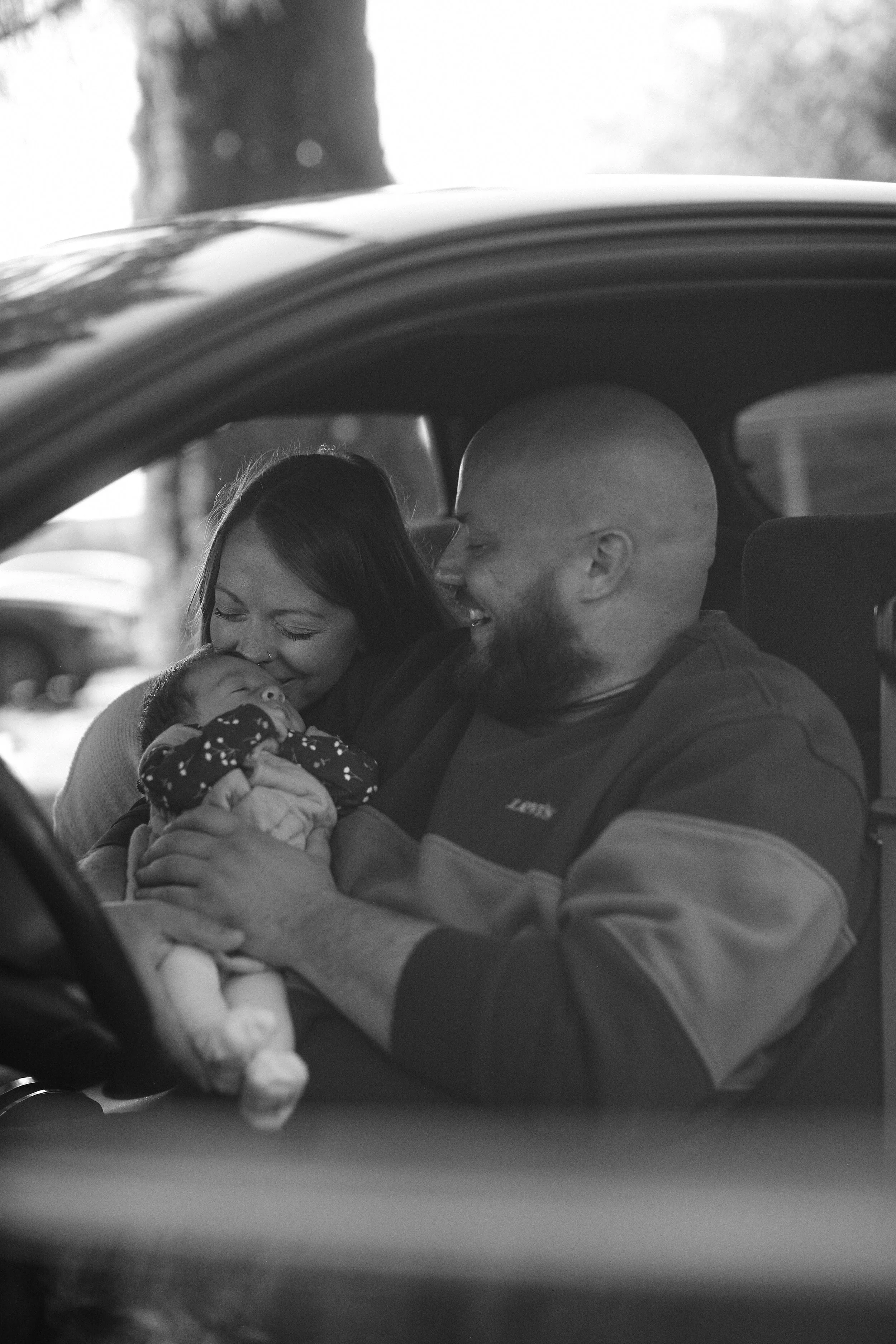 A black and white photo of a couple sitting in a car, smiling and holding a baby.