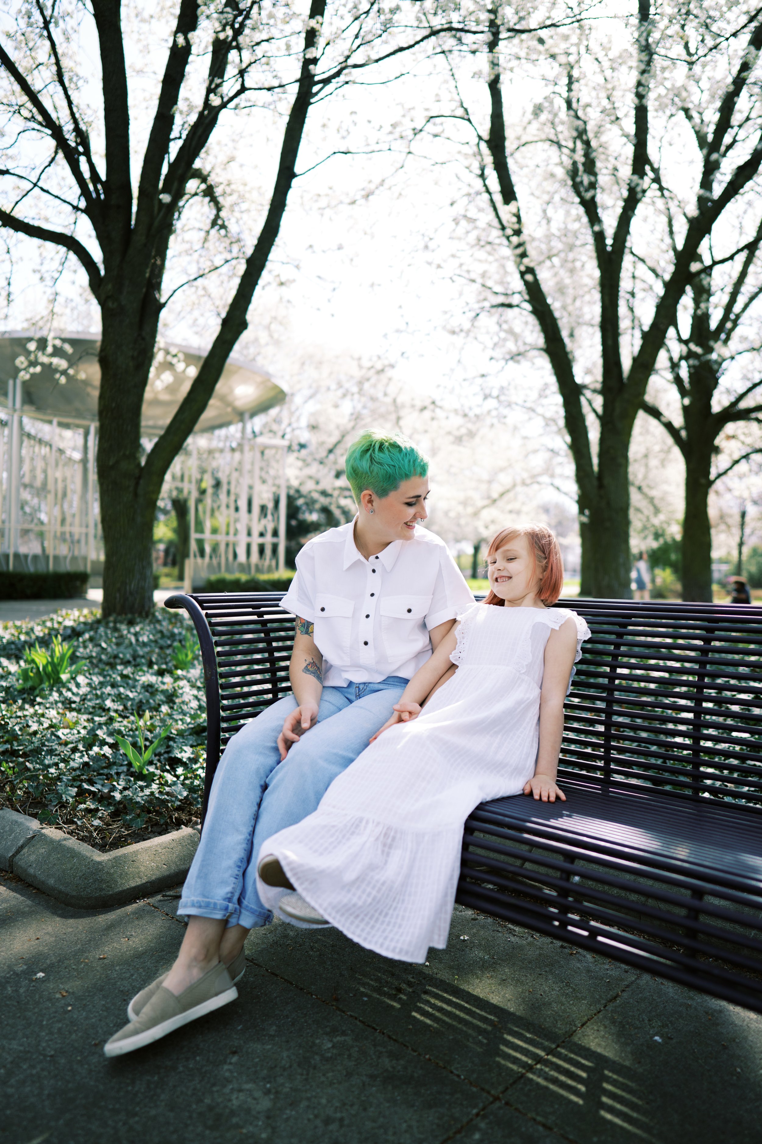 A person with green hair and a short-sleeve white shirt sits on a park bench with a young girl in a white dress. They are both smiling at each other. The background features blooming trees with white flowers.