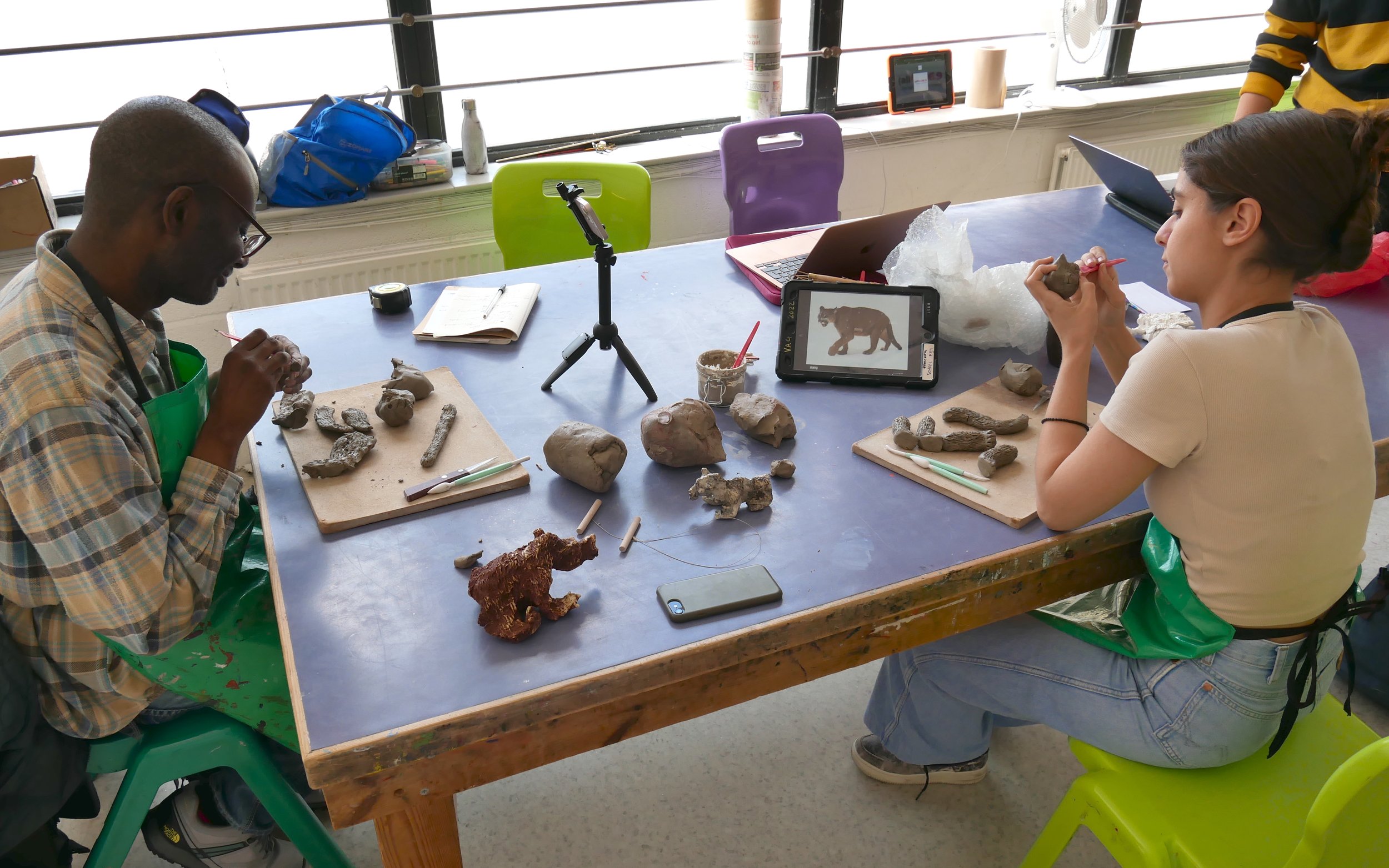  Leslie and Eleni working on clay animal sculptures together in the Venture Arts studio. Leslie is on the left and Eleni is on the right, both working on wooden boards. The photos show the animals in varying places along the journey of becoming full 