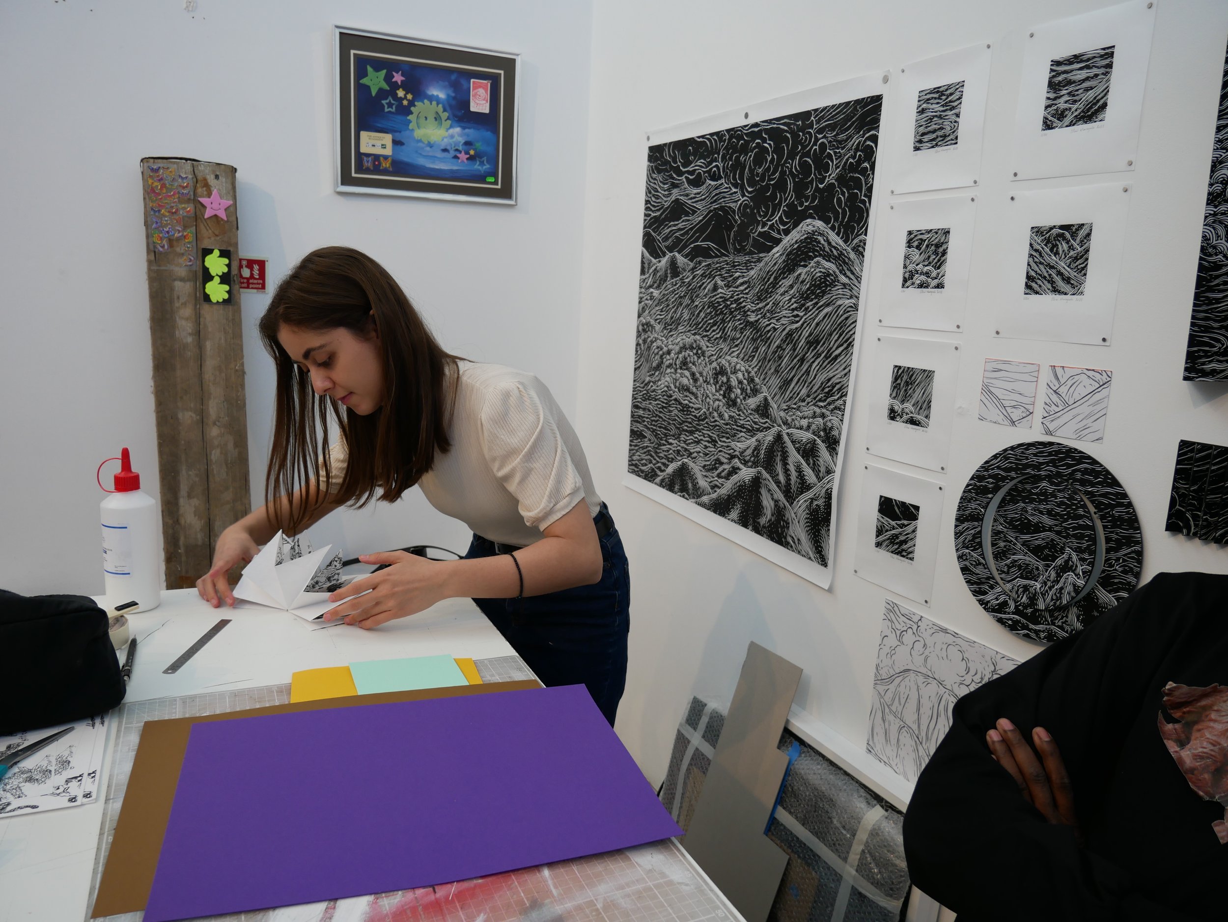  This series of photographs shows Eleni and Leslie creating folded books together in Leslie’s studio. Eleni is in a beige top and dark jeans, and Leslie is in a black top with a printed picture of a brown bear hanging around his neck. Artwork feature