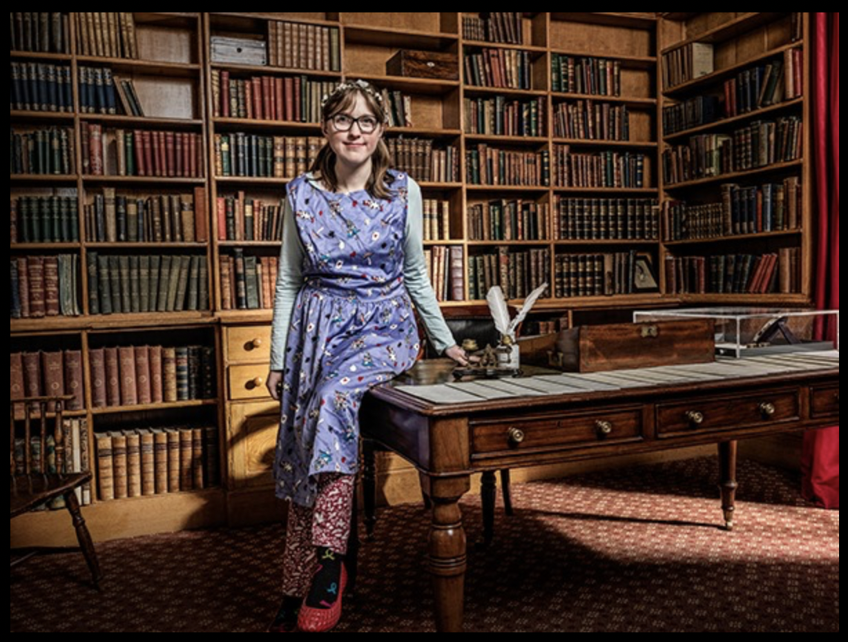 A photo of a young white woman in a patterened dress over patterned trousers, sat perched on a table infront of a very dense bookcase