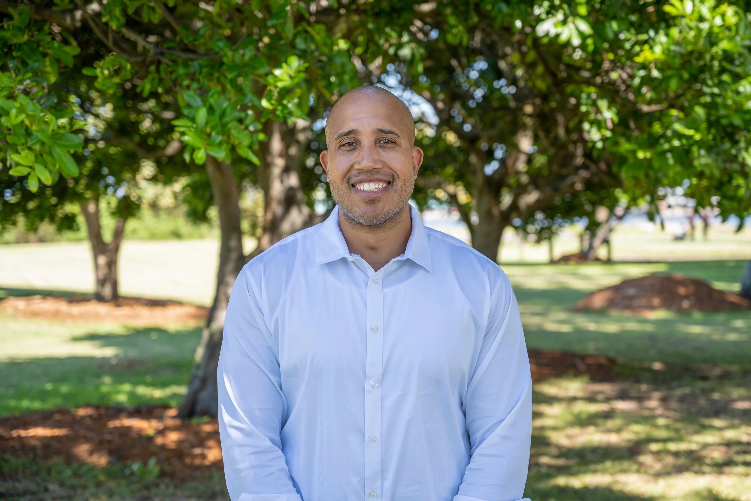 A smiling man in a white dress shirt standing outside in a park-like setting with green trees and grass in the background.