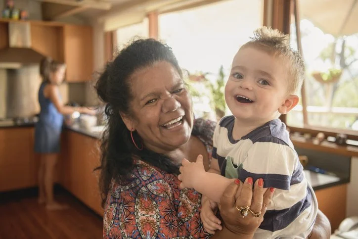 A joyful older woman holding a young boy in her arms inside a kitchen, with a girl in the background at the sink.