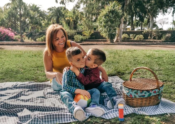 A woman and two young boys sitting on a picnic blanket outdoors, with the boys hugging each other. There is a picnic basket and some bottles nearby, and trees in the background.