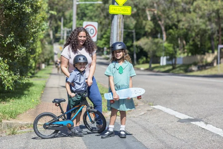 A woman and two children wearing helmets standing on a sidewalk near a bicycle and skateboard, smiling and enjoying a sunny day.
