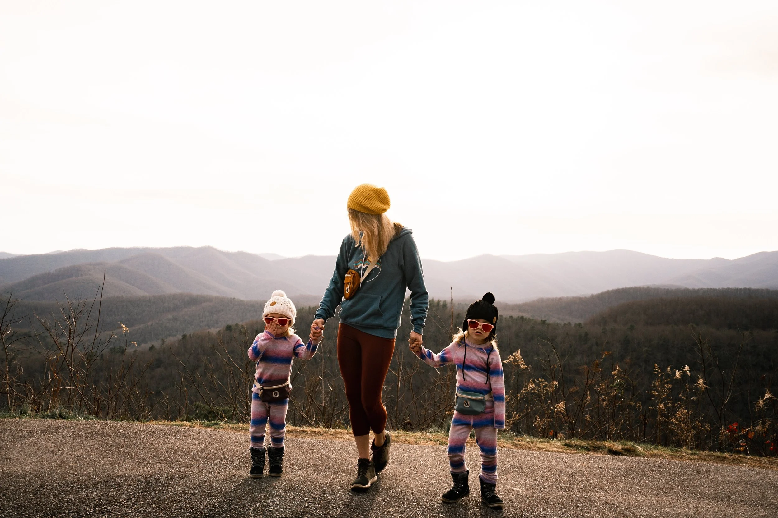 A woman walking outdoors with two young children, holding hands, in front of a mountain landscape during daytime, all wearing casual clothes and sunglasses.
