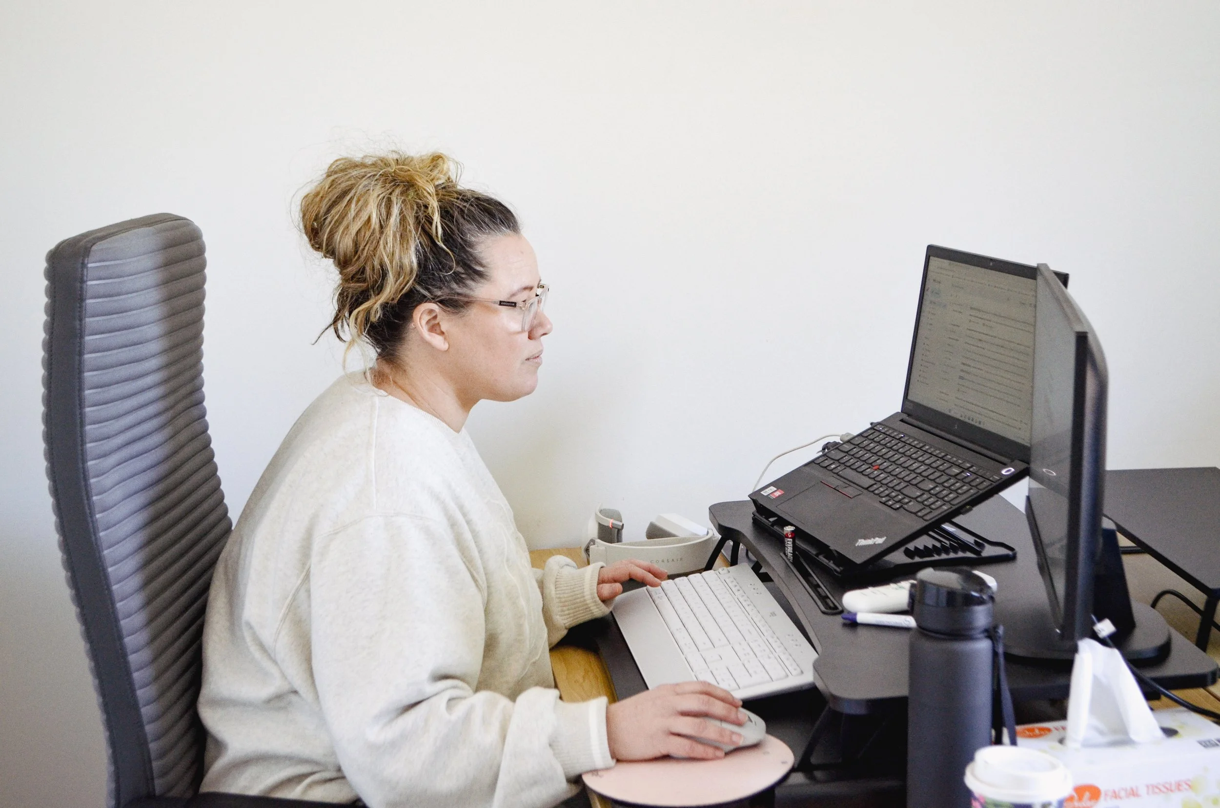A woman with glasses and curly hair in a messy bun working at a desk with a computer, multiple monitors, a keyboard, and office supplies.