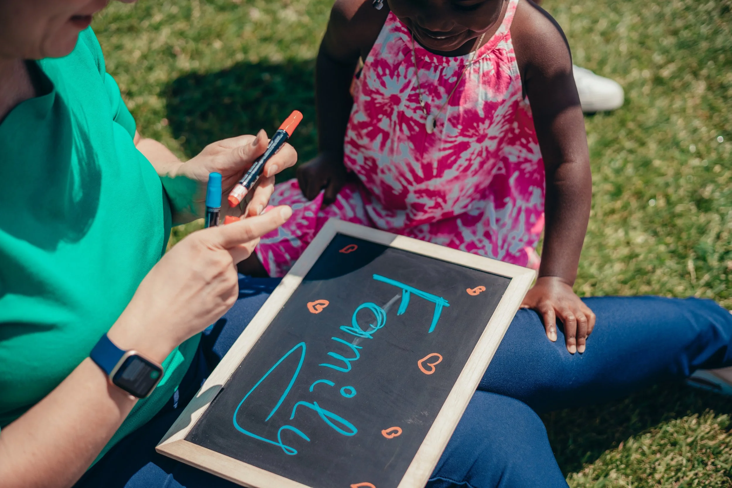 A person holding a chalkboard with the word 'TIME' written on it, sitting outdoors on grass, with a young girl looking at the chalkboard and a person next to her pointing at it. The girl is wearing a pink and white dress, and the scene takes place on a sunny day.
