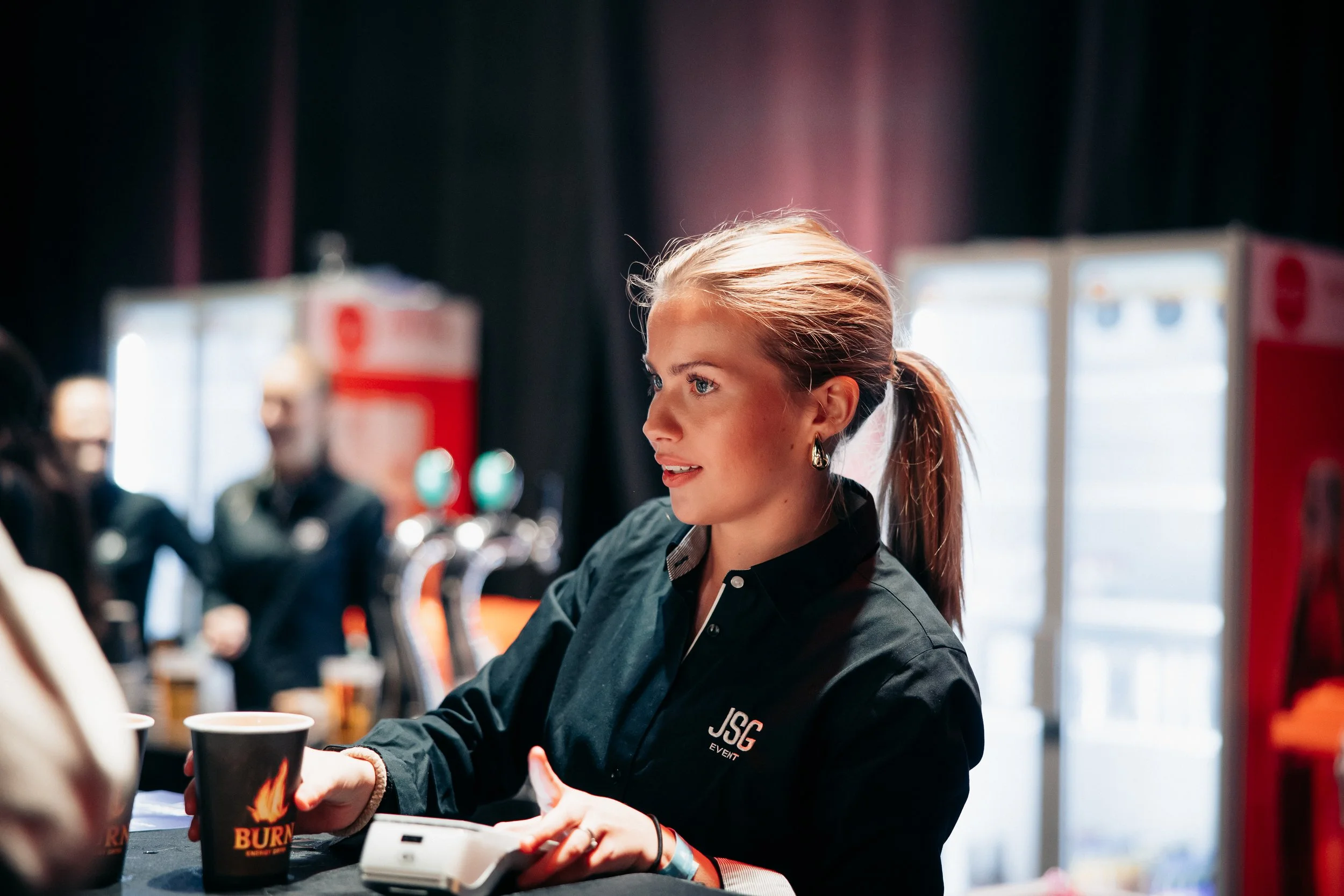 En kvinne med blond hestehale og svart T-skjorte med logo som står i en eventbar og rekker en svart kopp til en kunde, med kjøleskap i bakgrunnen.