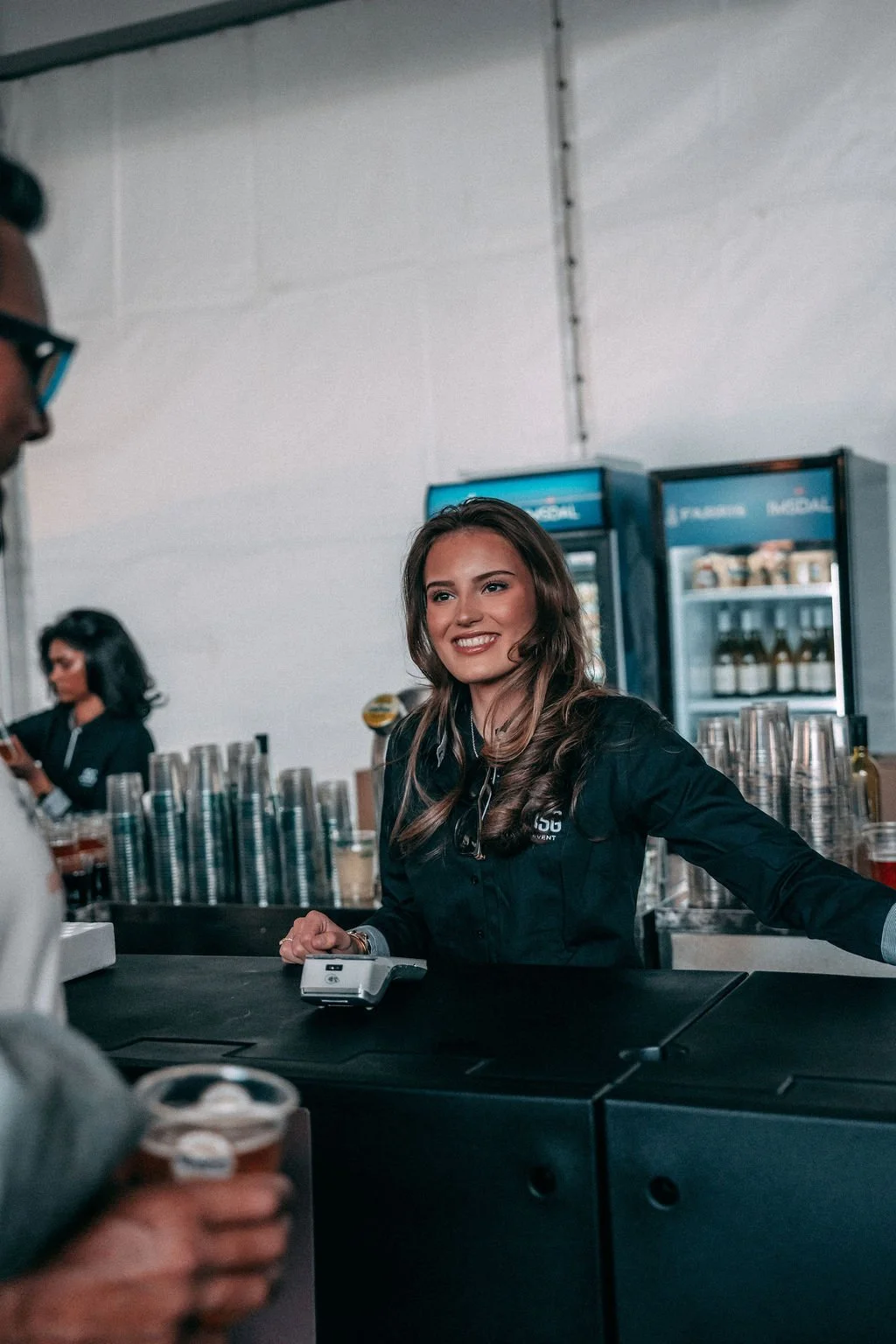 En smilende kvinnelig festivalansatt med langt brunt hår som serverer en kunde i en festivalbar, med barområde og annet crew i bakgrunnen.