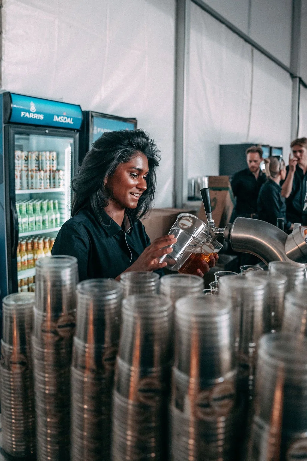 En kvinne i svart T-skjorte som tapper øl i en festivalbar, med drikkekjøleskap og festivaldeltakere i bakgrunnen.