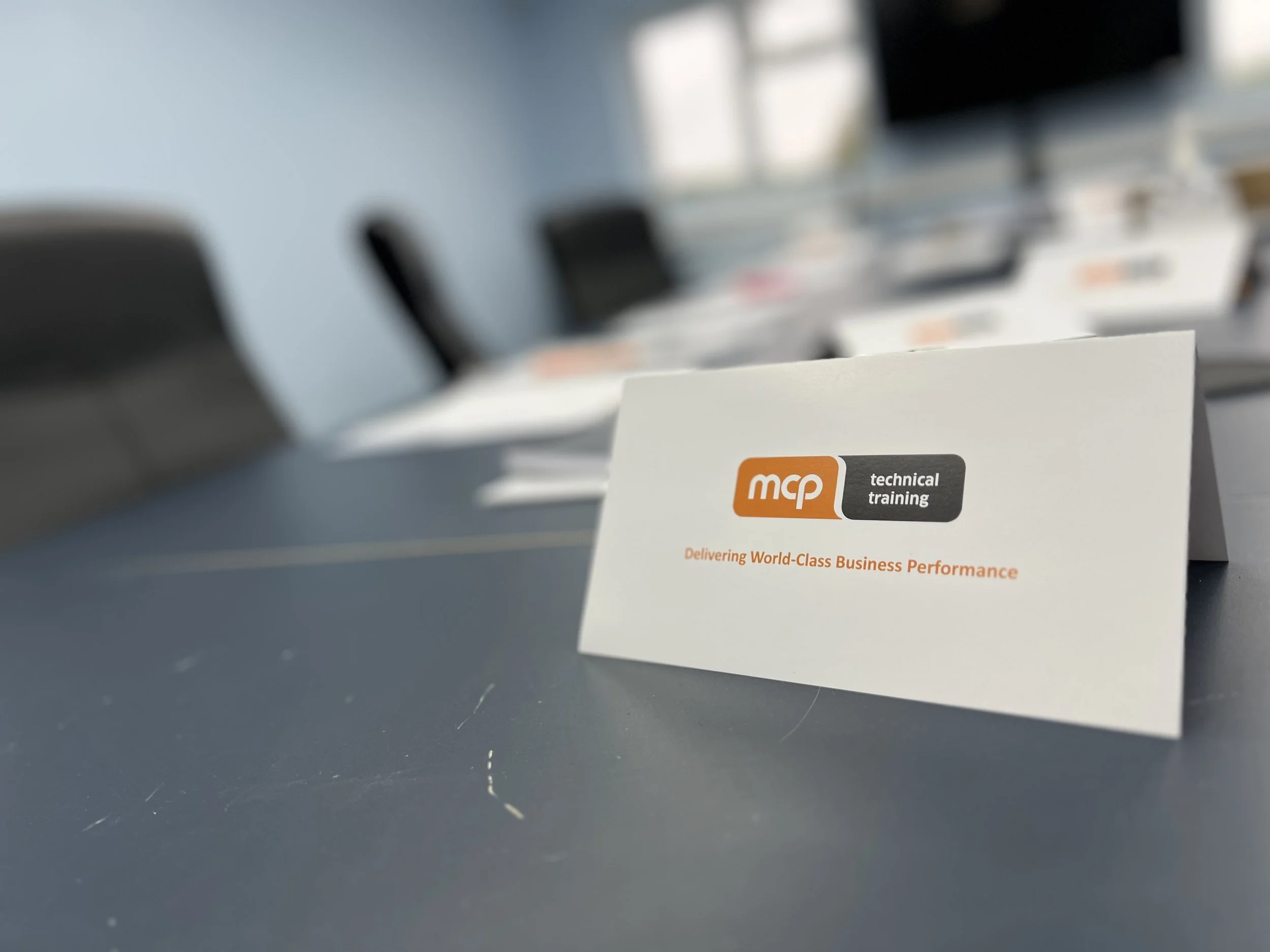 A white card on a conference table with the mcq technical training logo and the slogan, 'Delivering World-Class Business Performance.' In the background, blurred conference chairs and tables are visible.