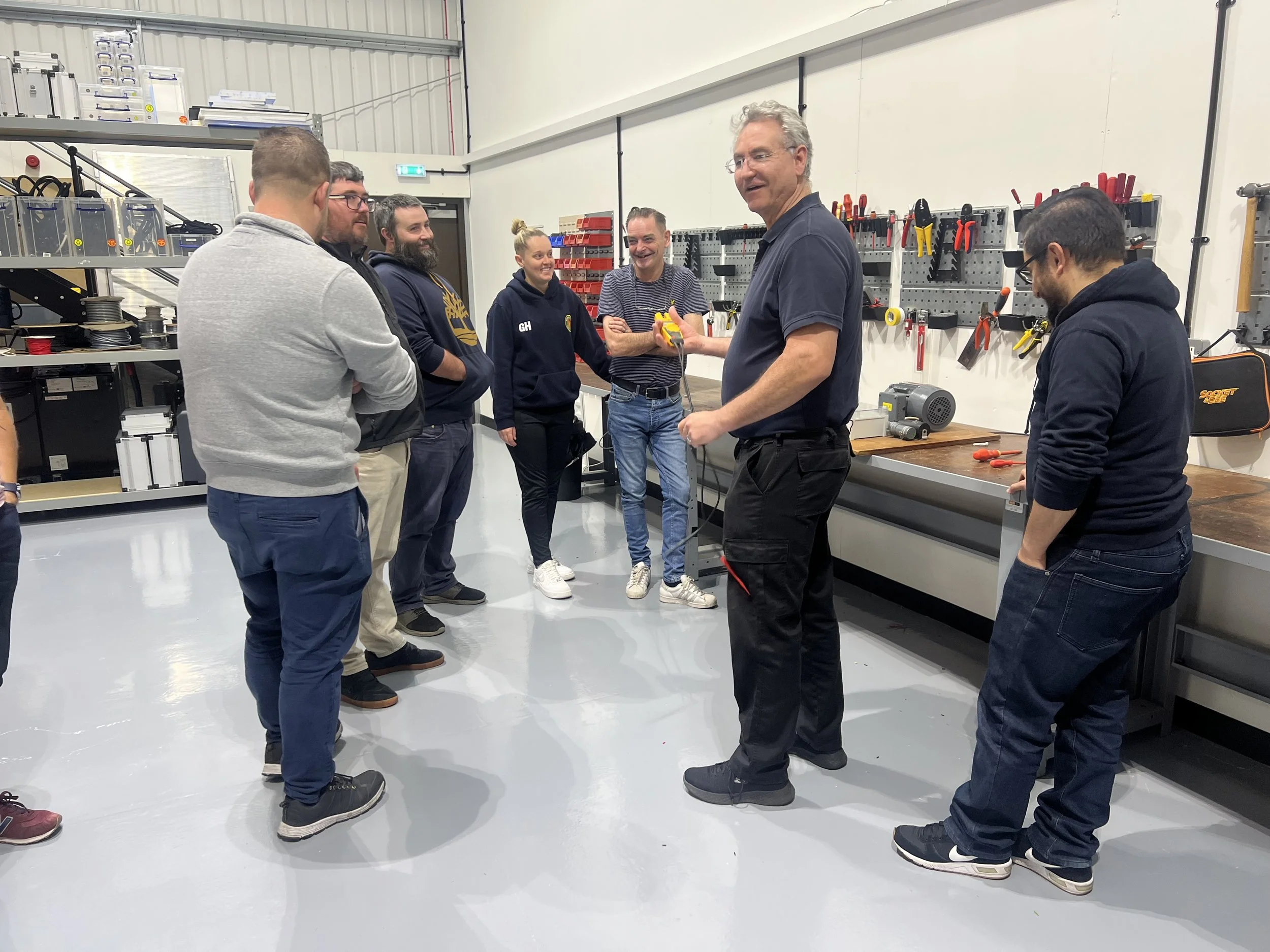 A group of seven people in a workshop, with tools and equipment on the wall, engaged in a discussion or training session.