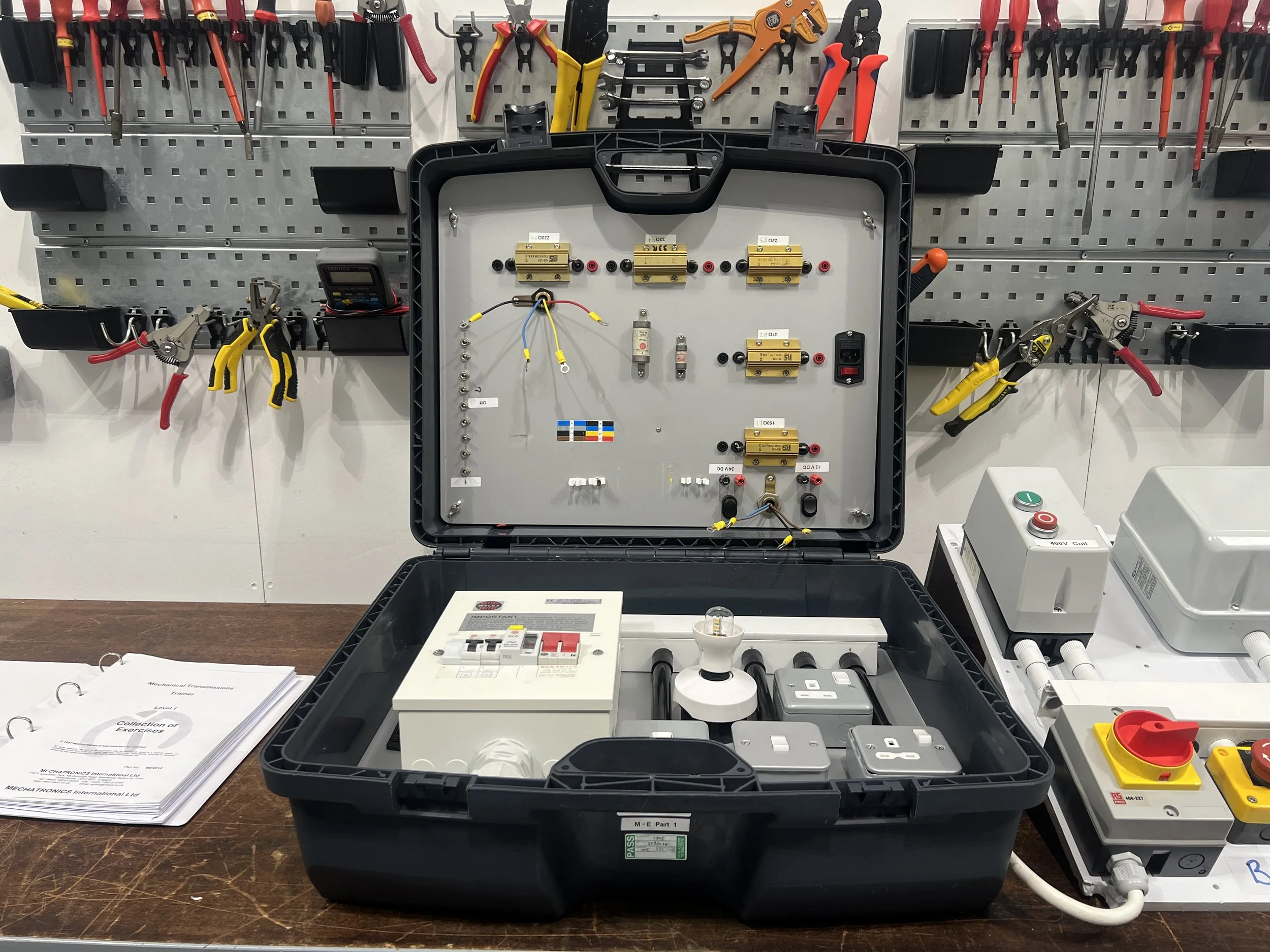An electrical training kit with various electrical components and tools arranged inside a black case, on a workbench with a pegboard holding tools and a sheet of paper.
