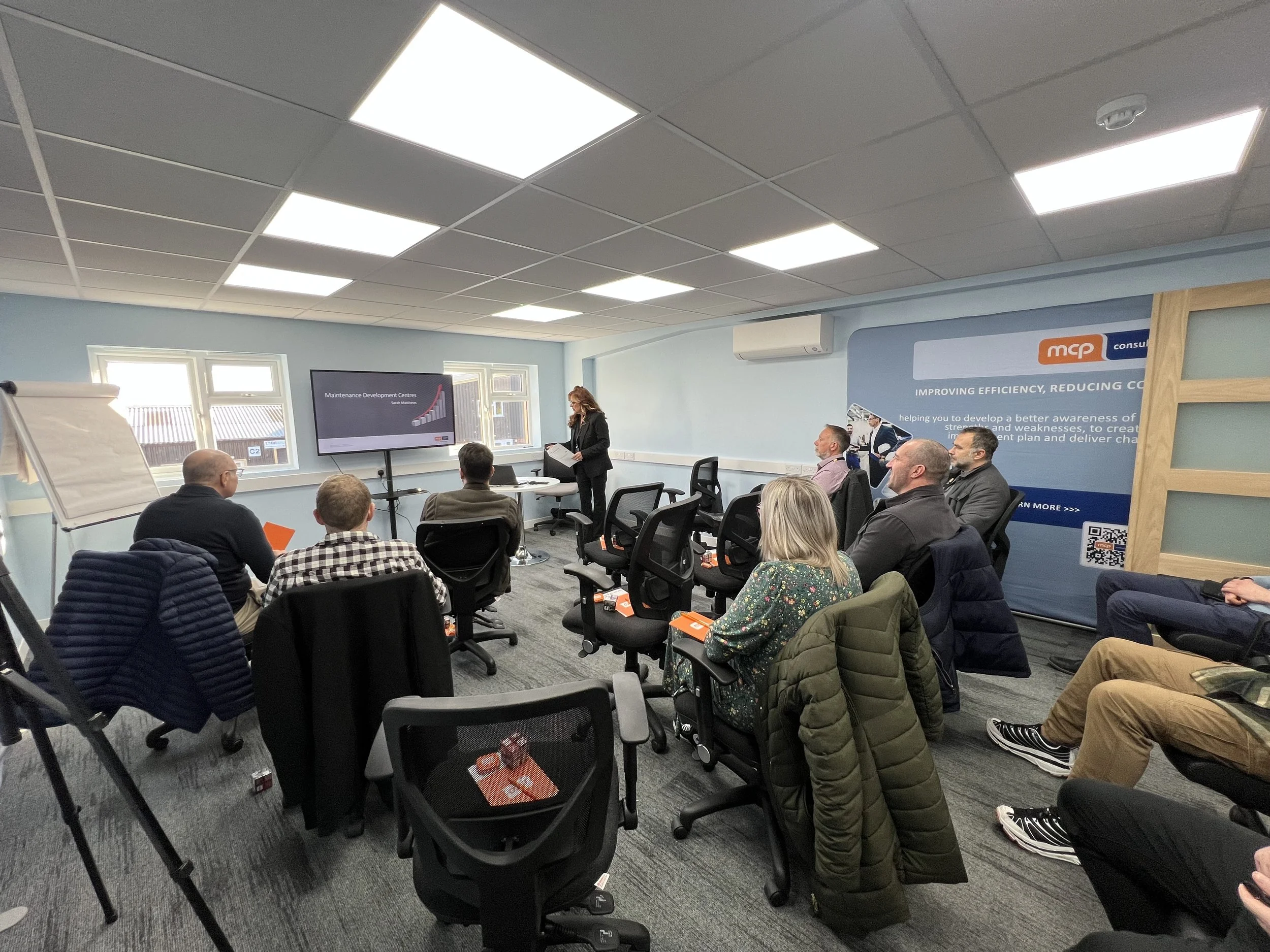 A business meeting or workshop taking place in a conference room with attendees seated facing a presenter standing at the front near a screen, while others listen and take notes.
