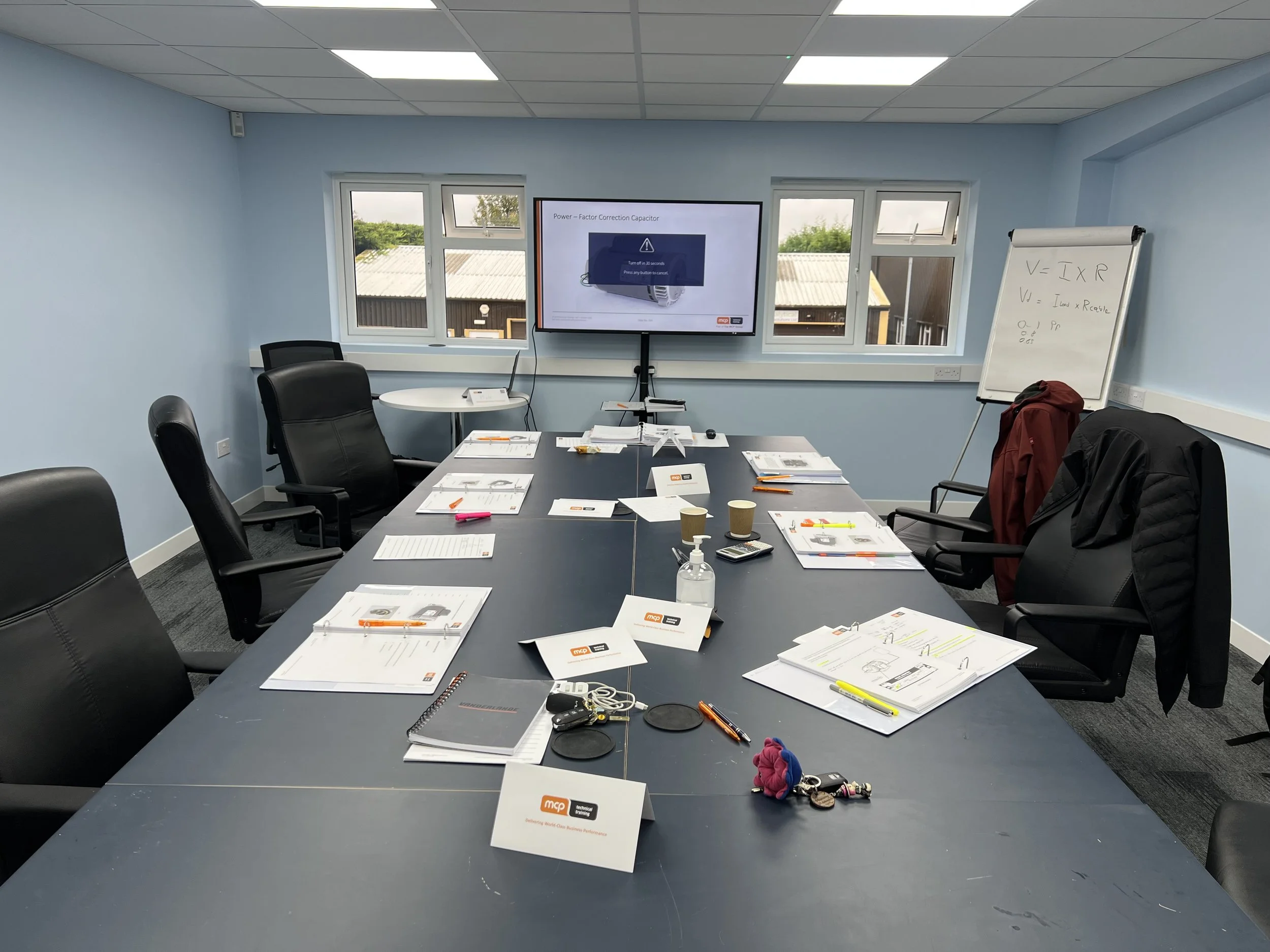 Empty conference room with a long table, black chairs, notebooks, pens, and a whiteboard, TV screen displaying a presentation, and two windows showing outdoor view.