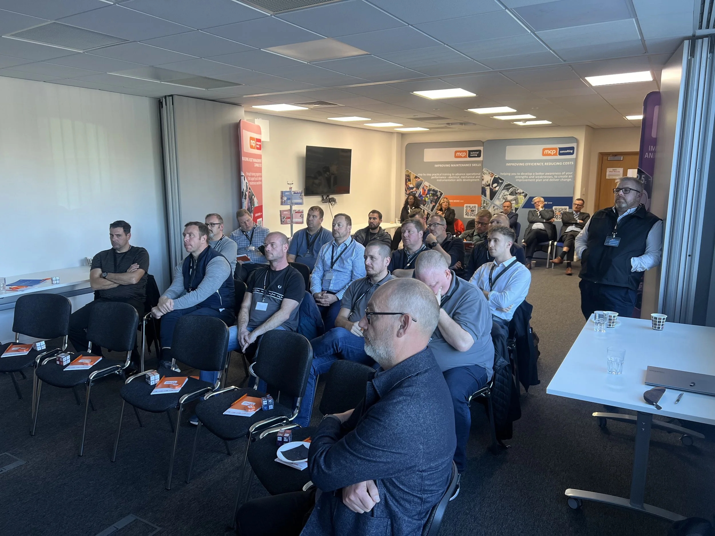 A group of men attending a presentation or conference in a conference room, some seated and some standing, with banners and a TV screen in the background.