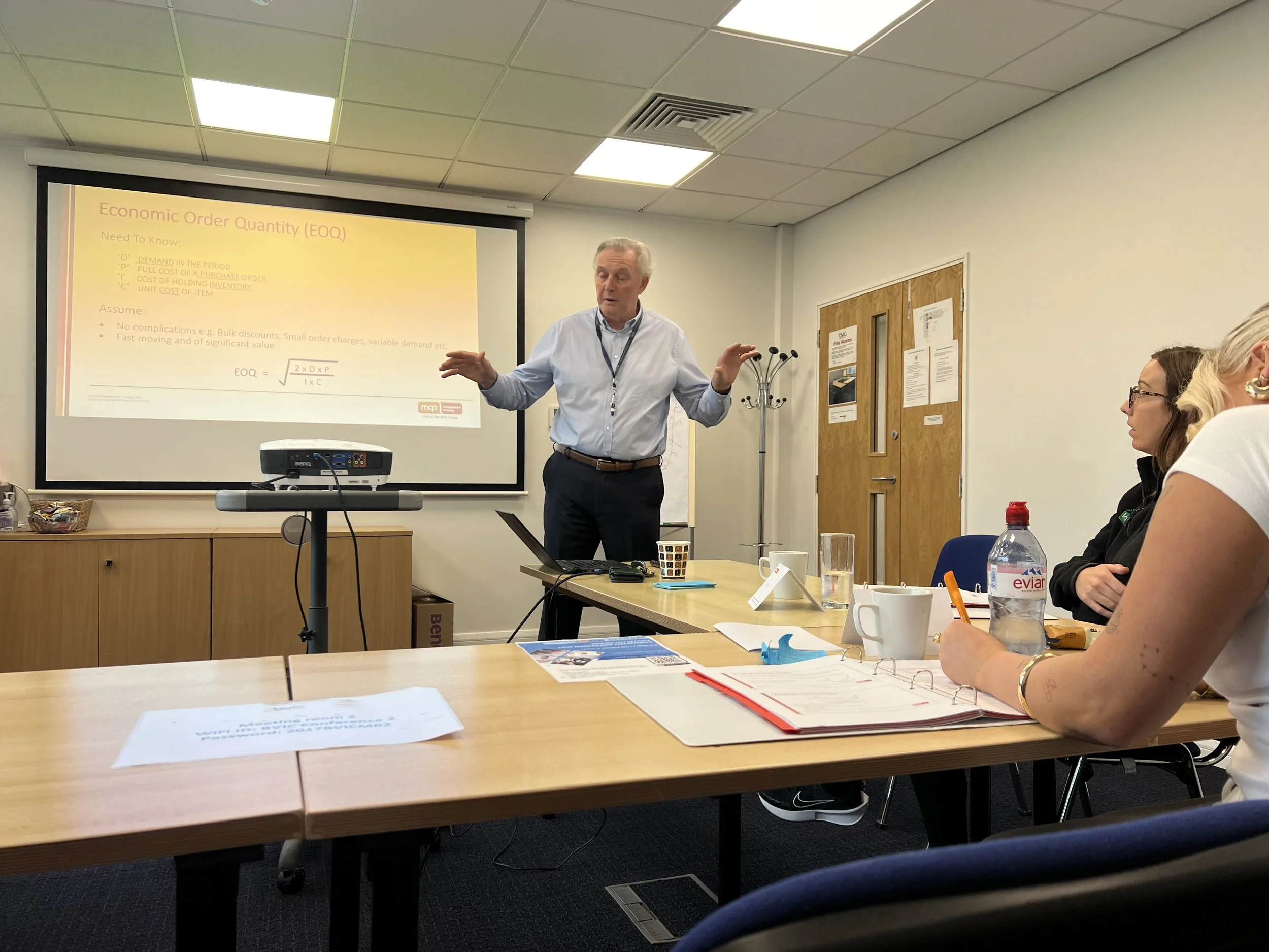 A man giving a presentation in a conference room with a slide on the screen titled 'Economic Order Quantity (EOQ)' and an audience of women seated at tables with notebooks, water bottles, and cups.