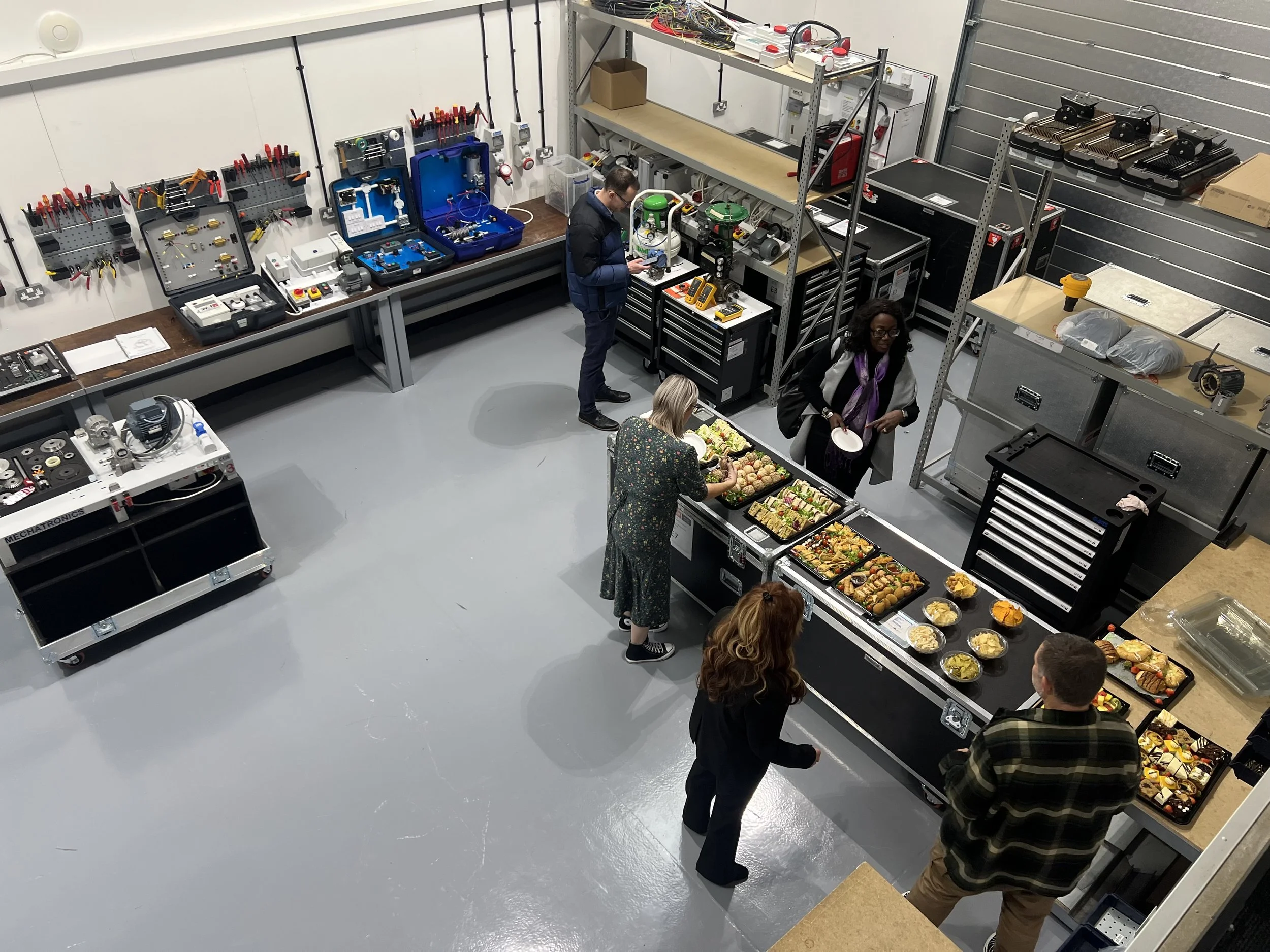 People at a buffet of salads and snacks in an industrial workshop or warehouse with tools, equipment, and electronics on shelves.