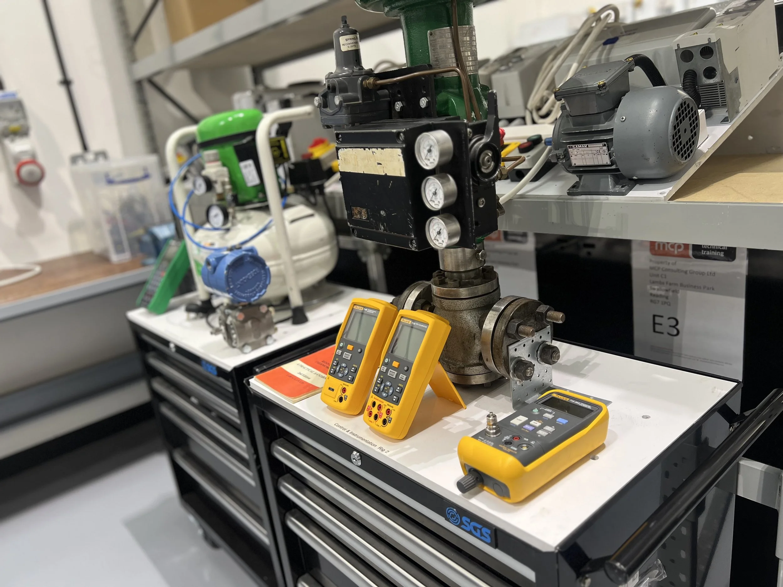 Industrial workshop setup with various tools and equipment, including two yellow digital multimeters and a portable clamp meter on a black tool cart, and mechanical components on shelves in the background.