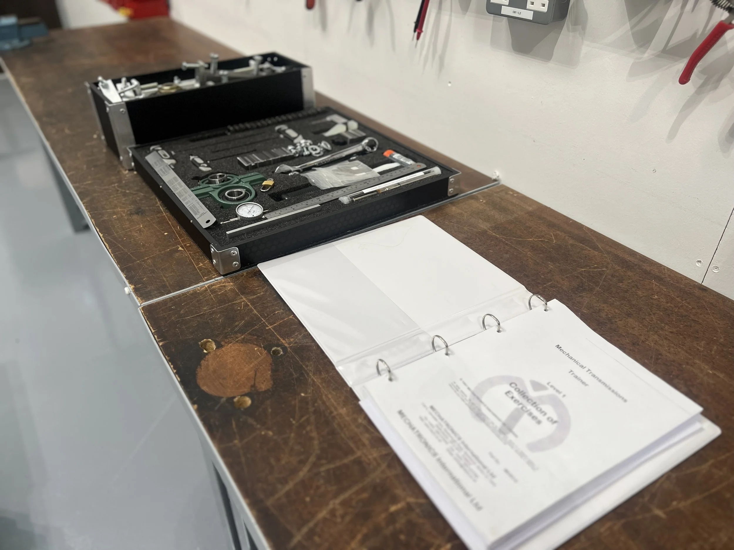 Open binder with training manual titled 'Collection of Exercises' next to a toolbox with various tools on a workbench.