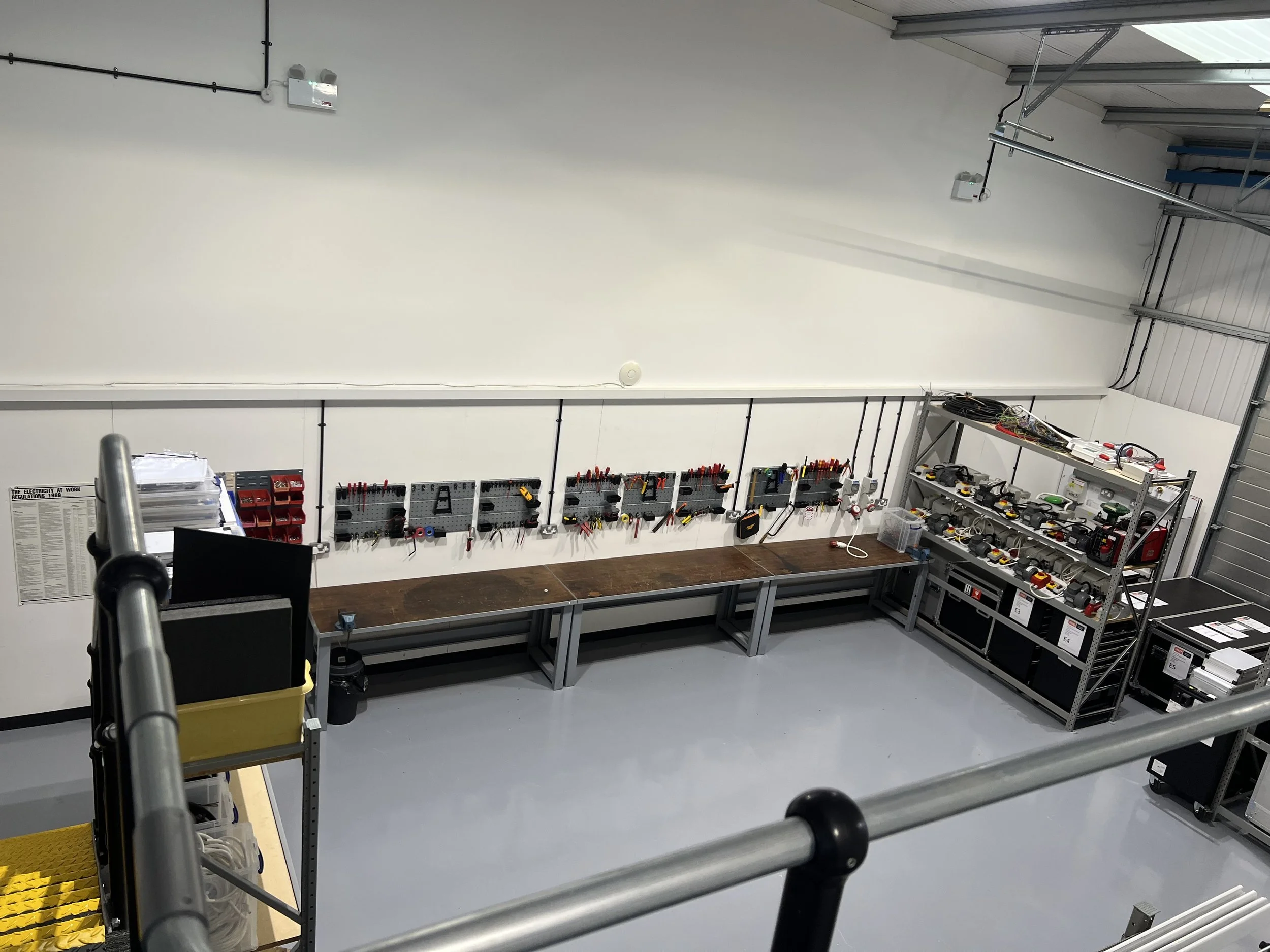 View of a clean, organized workshop or laboratory with a long workbench, pegboards with tools, shelving units with various electronic tools and equipment, and a rolling cart in the foreground.