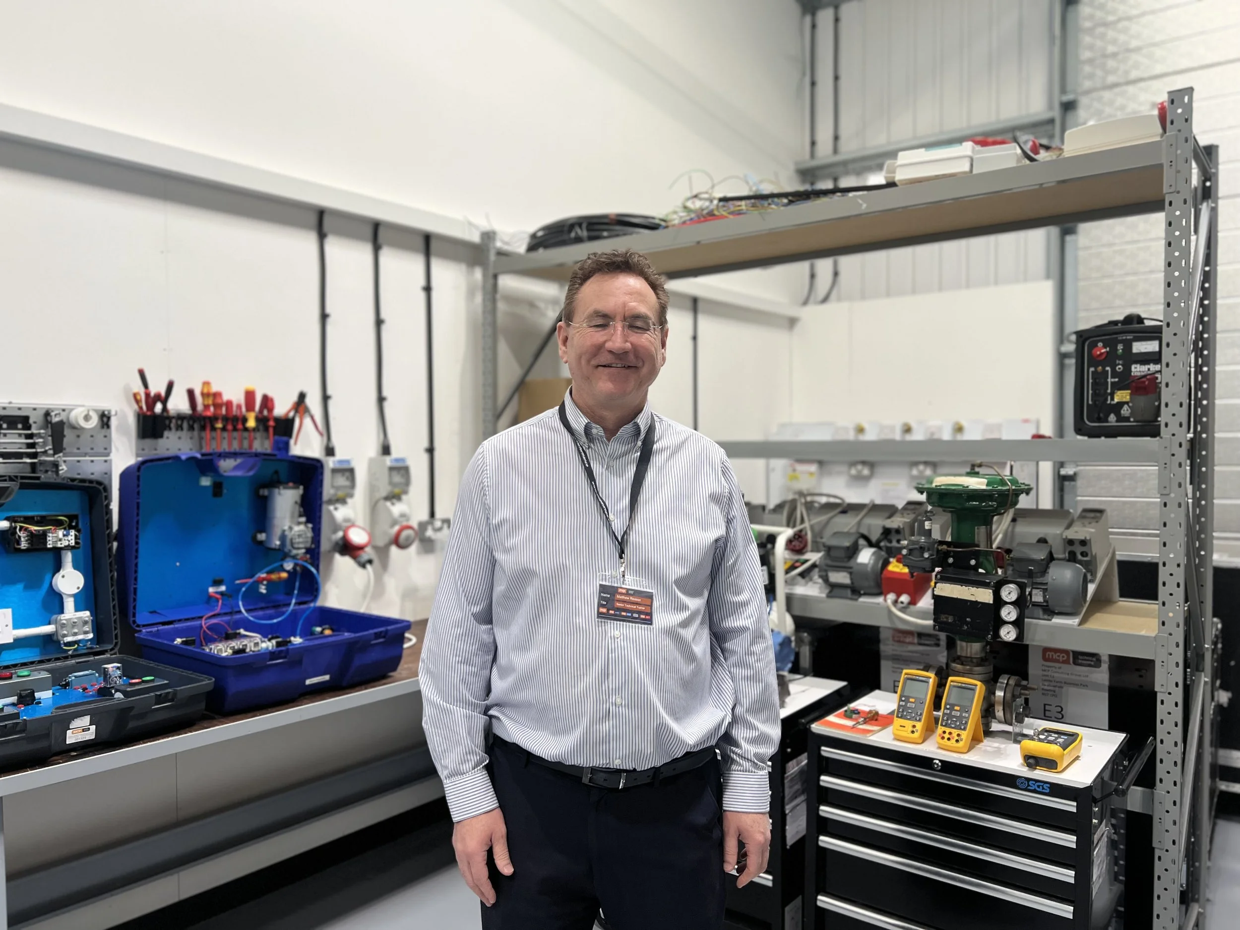 A man in business attire smiling in an industrial workshop with electronic and mechanical equipment, tools, and multimeters on shelves and workbenches.