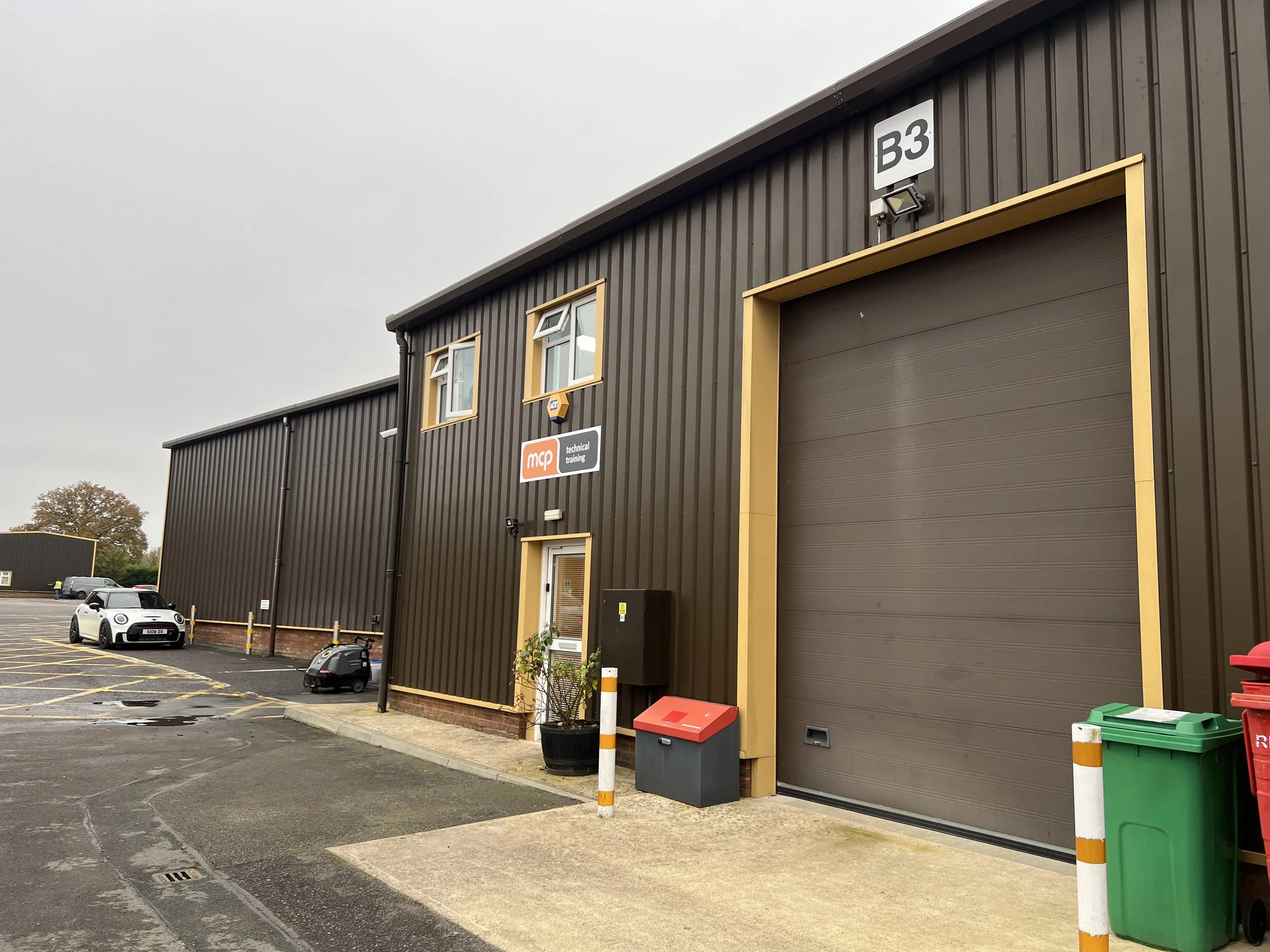 Industrial building with a large garage door, small windows, and parking lot in front, on an overcast day.