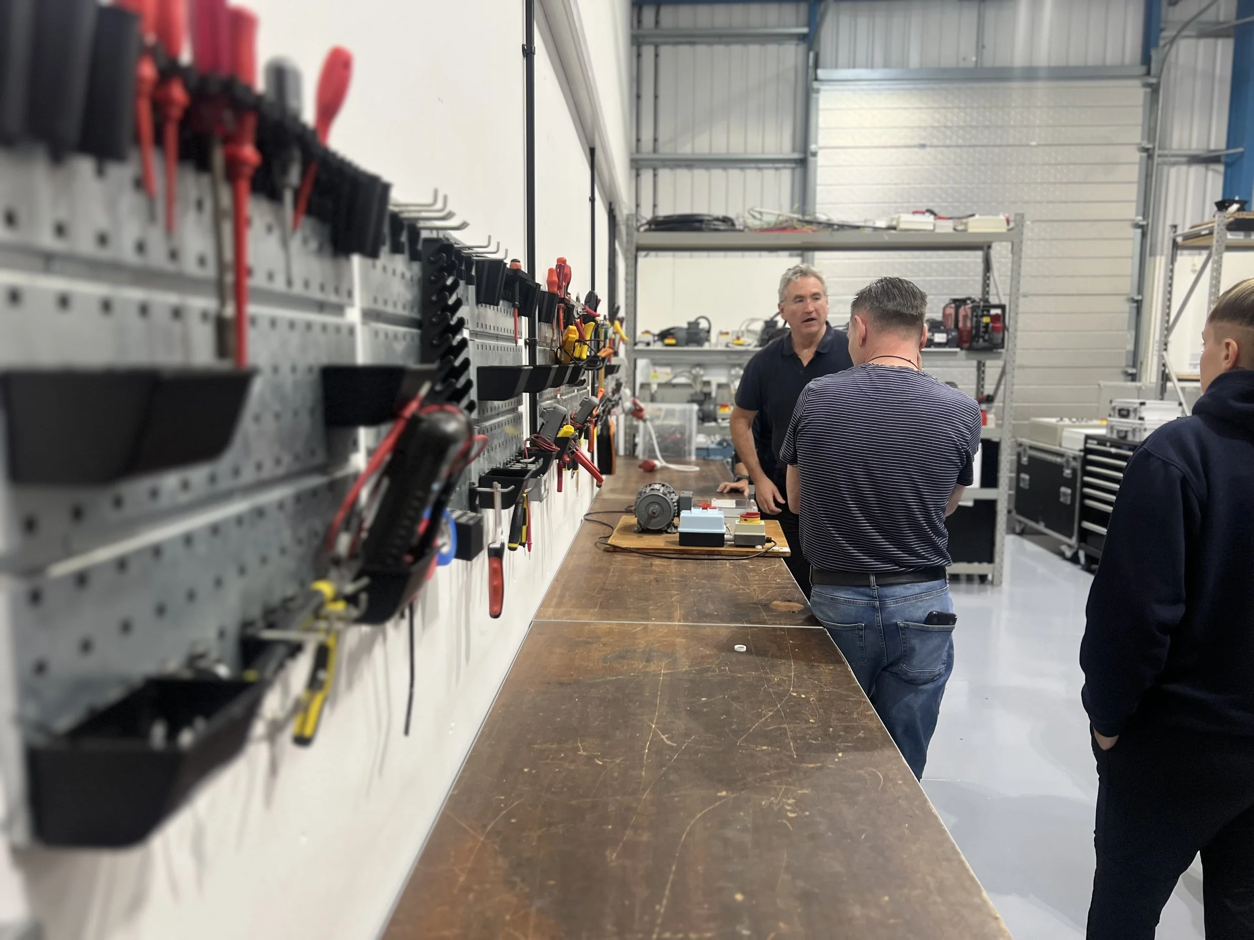 Three men are in a workshop, one is standing and talking while the other two listen. The workspace has tools hanging on a pegboard and workbenches with equipment.