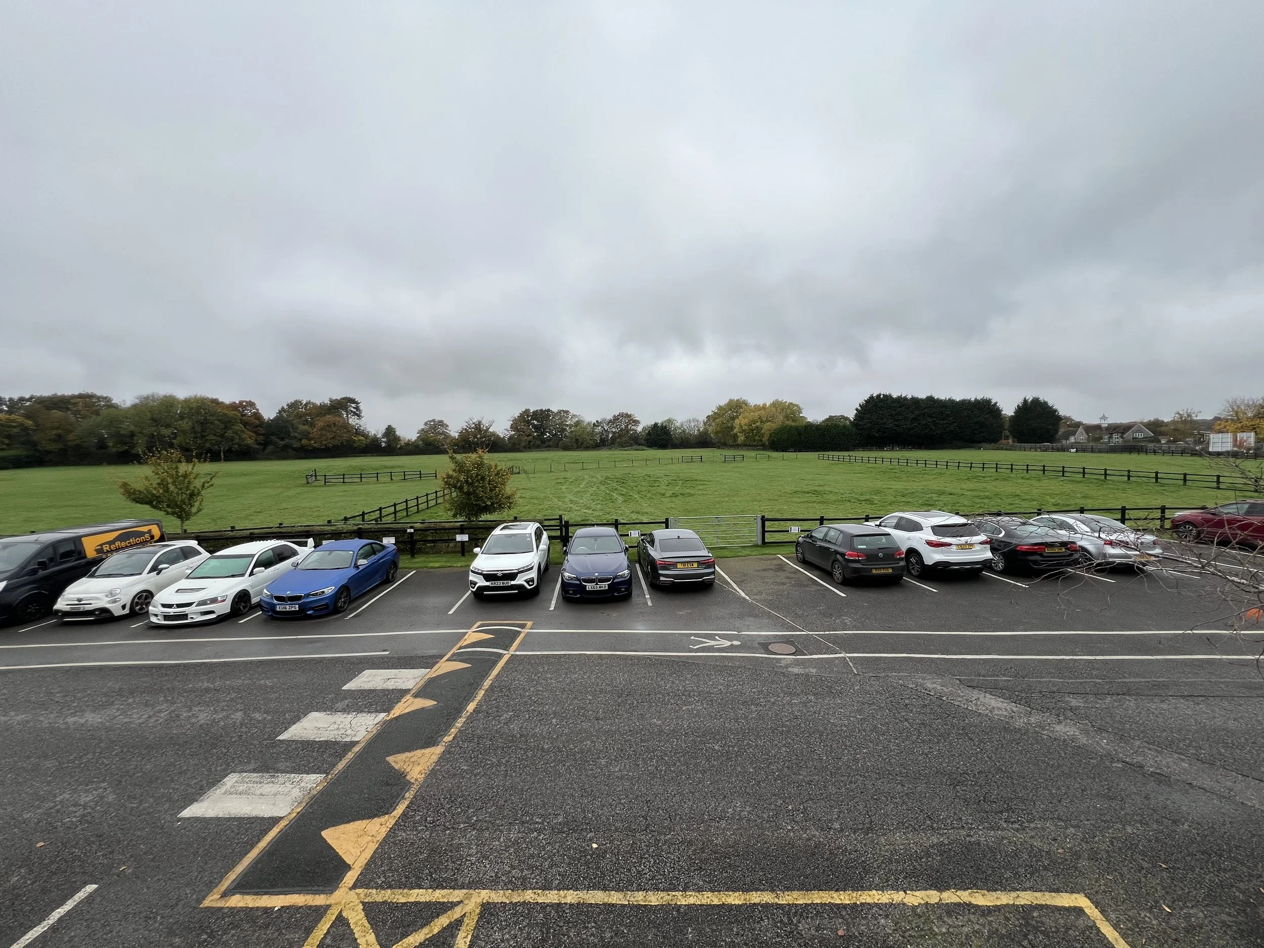 Parking lot with cars parked along a green grassy field, trees and overcast sky in the background.