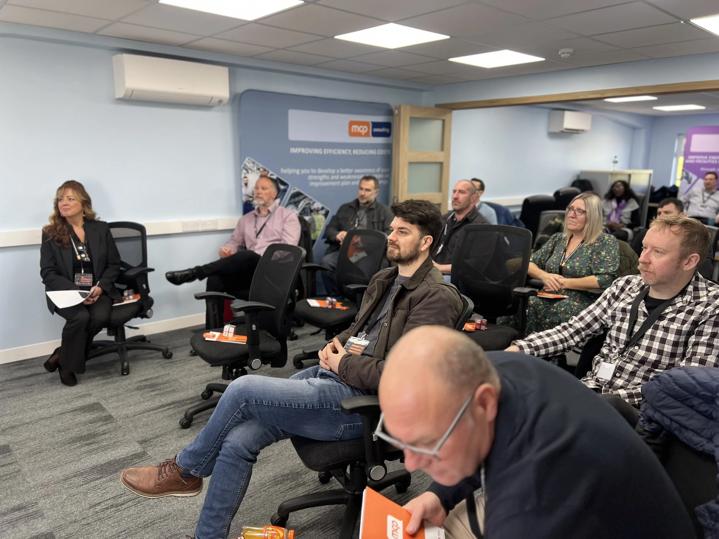 A group of people attending a conference in a room with light blue walls, some listening attentively while others are taking notes or looking at their devices.