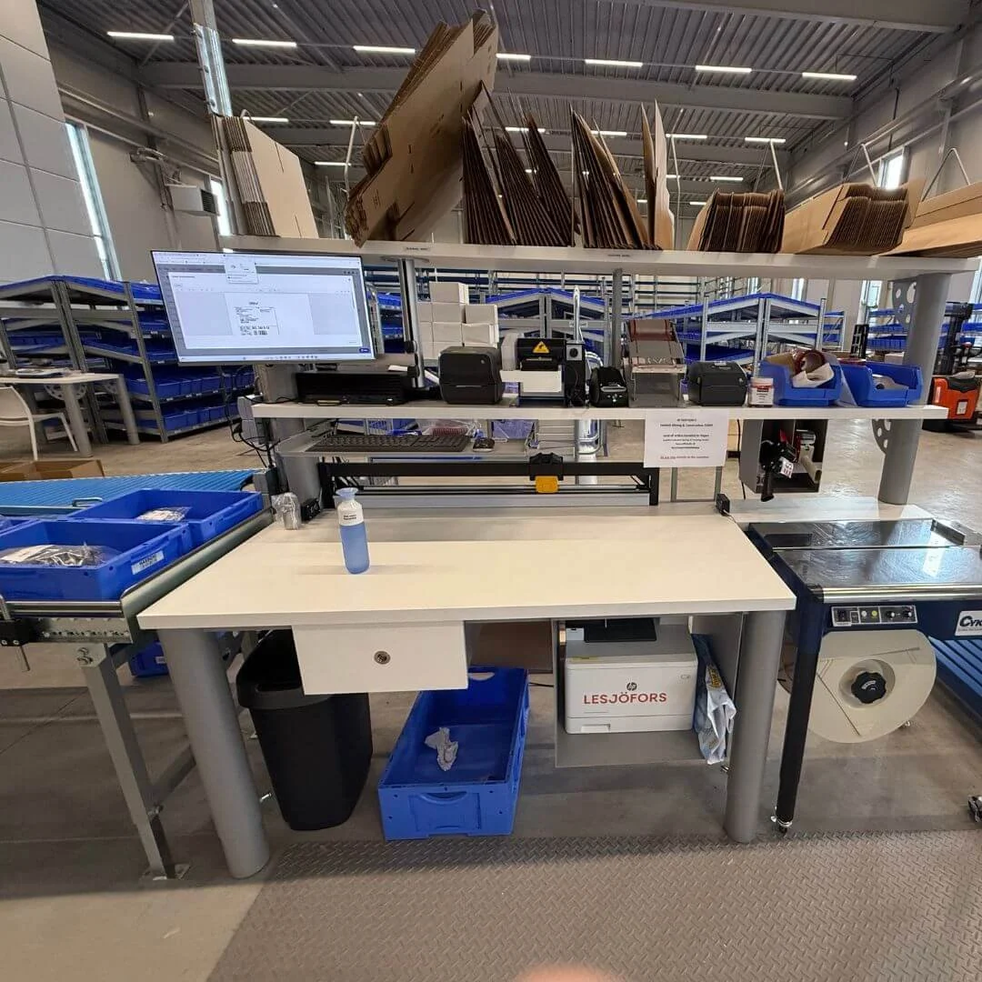 Packstation i Lesjöfors centrallager med automationsutrustning, skärmar, etikettskrivare och blå backar för orderplock.