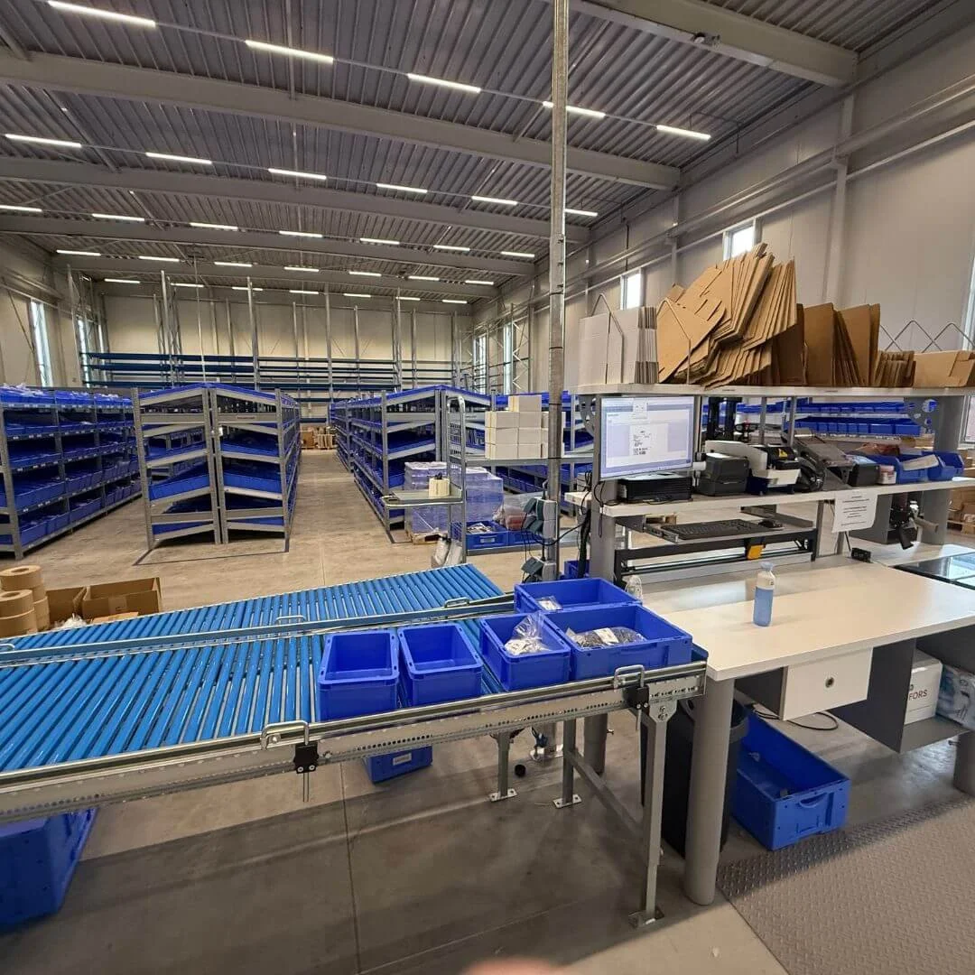 Plockstation och transportband i Lesjöfors centrallager i Nederländerna, med blå plastbackar och digitalt packflöde.