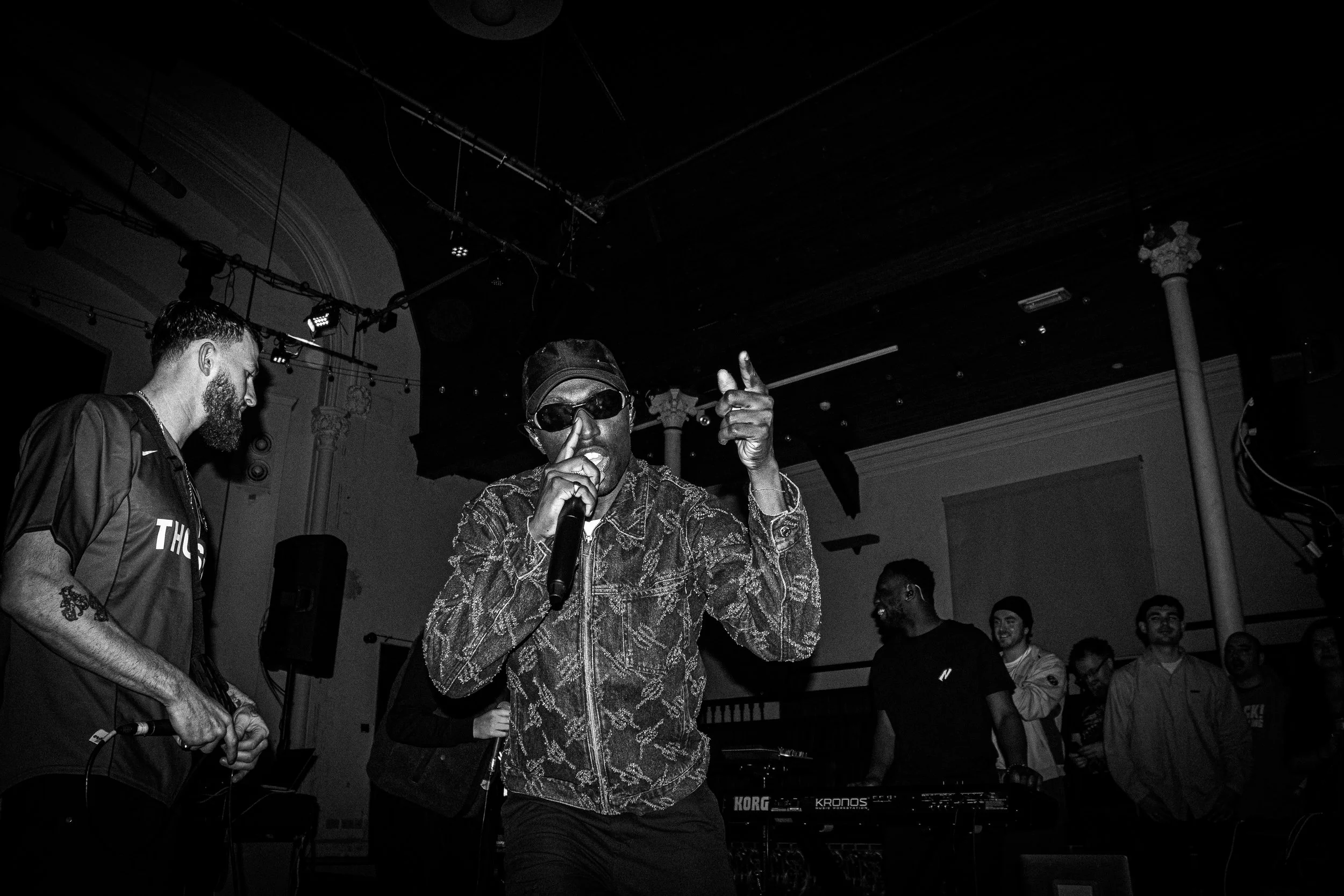 A black-and-white photo of a performer on stage wearing sunglasses and a patterned jacket, holding a microphone, with a group of people smiling and watching in the background.