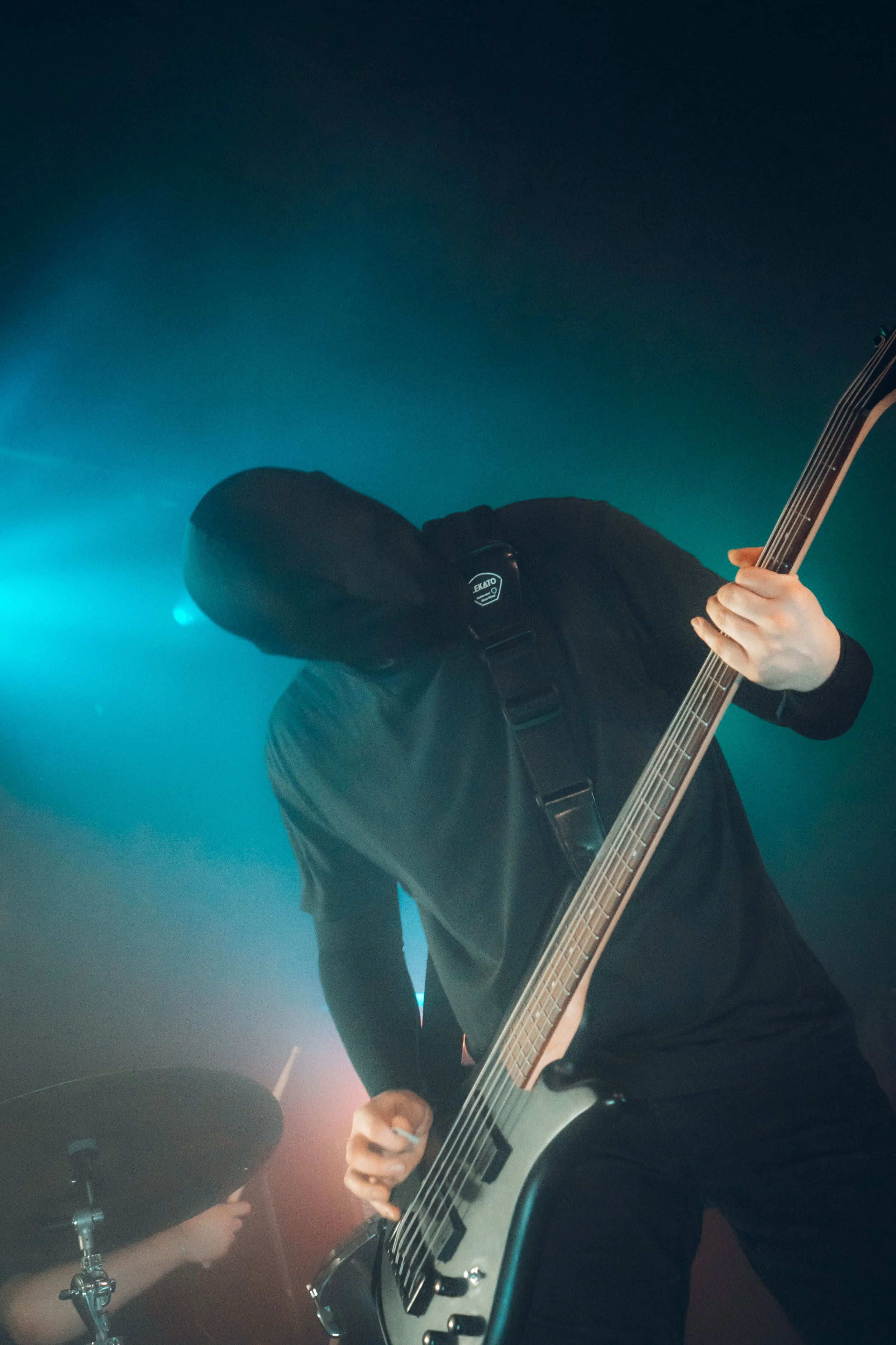 A person playing an electric guitar on a stage with blue and red lighting, and a drummer partially visible in the background.
