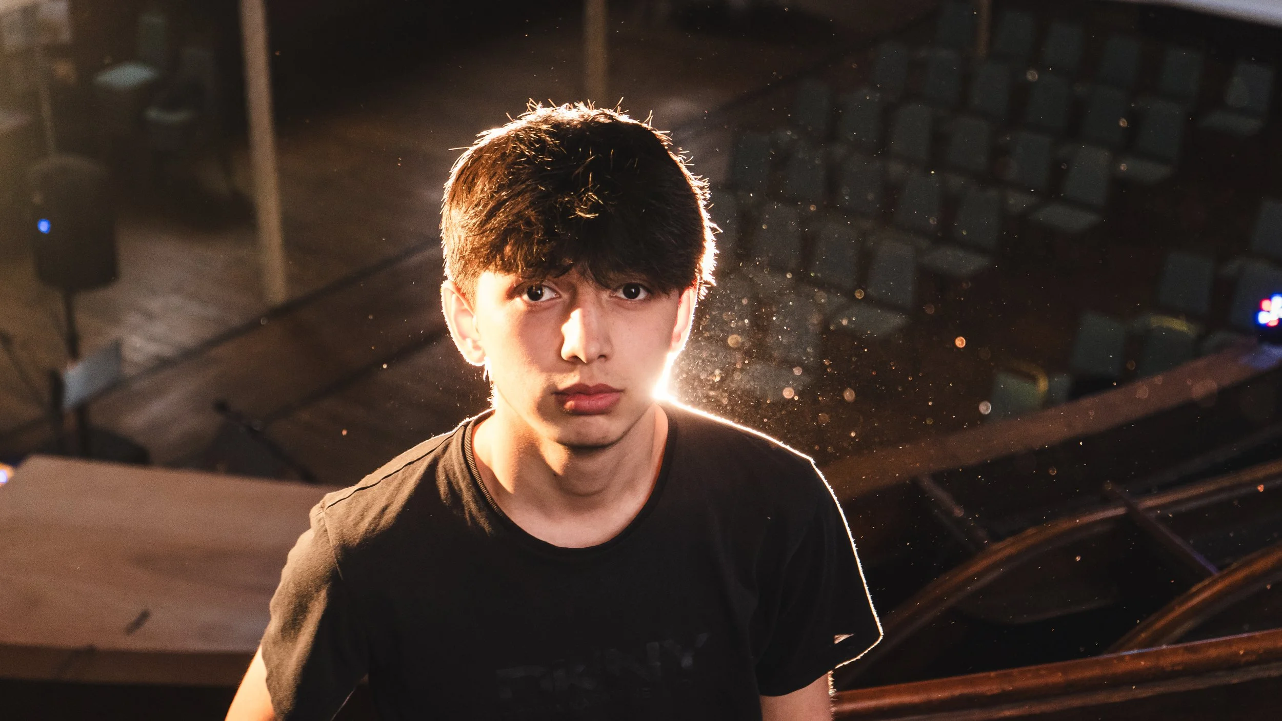 A young man with dark hair and a serious expression stands in a dimly lit environment, with bright backlighting highlighting his silhouette and dust particles in the air.