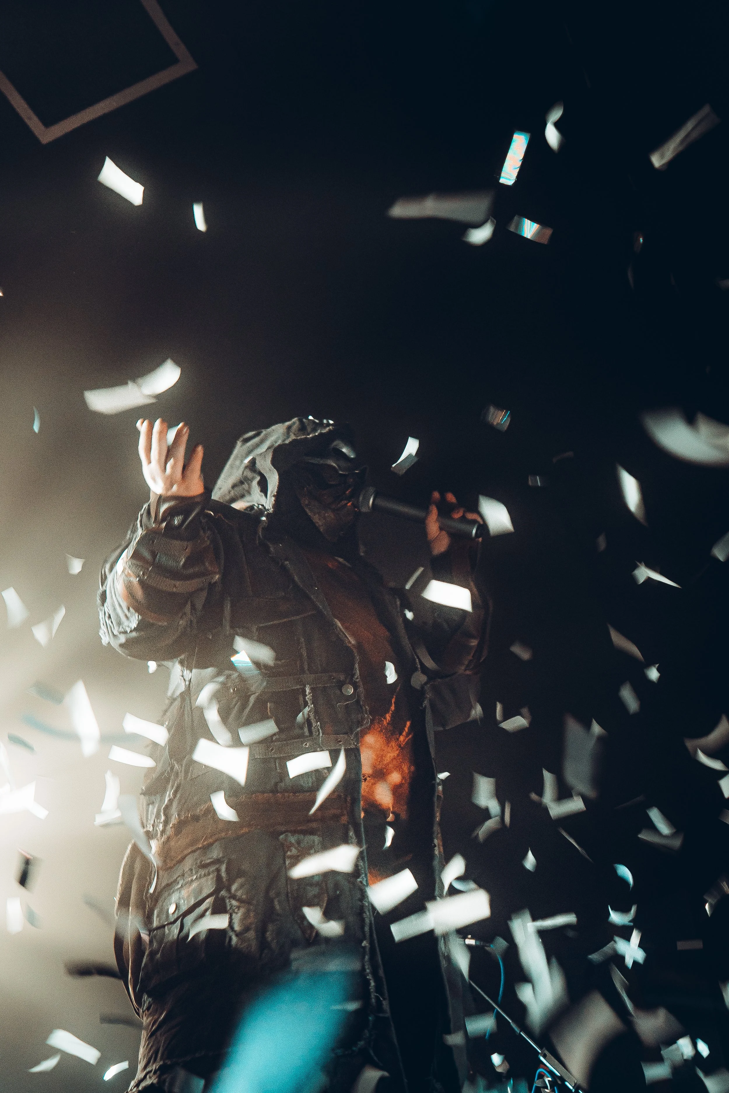 Person in a dark jacket and hood surrounded by falling white confetti in a dimly lit environment.
