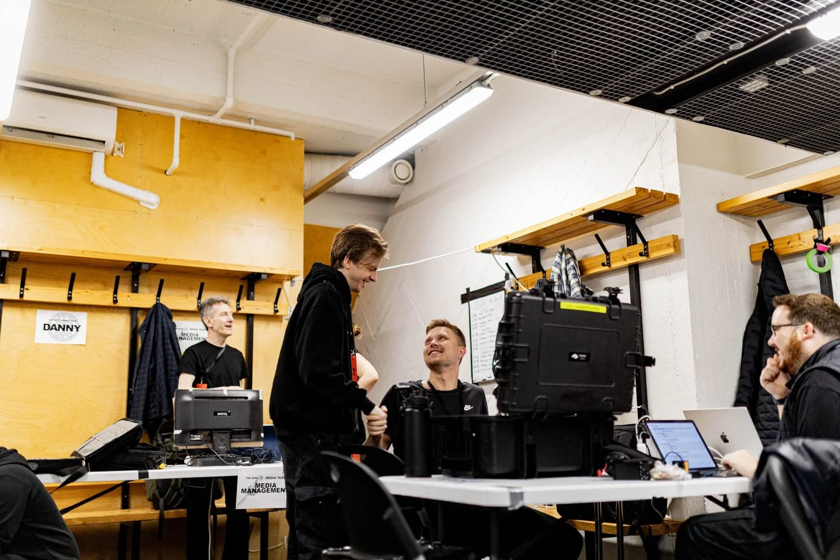 Group of people in a room with wooden and white walls, some talking and smiling, working at tables with laptops, cameras, and equipment, in a collaborative workspace.