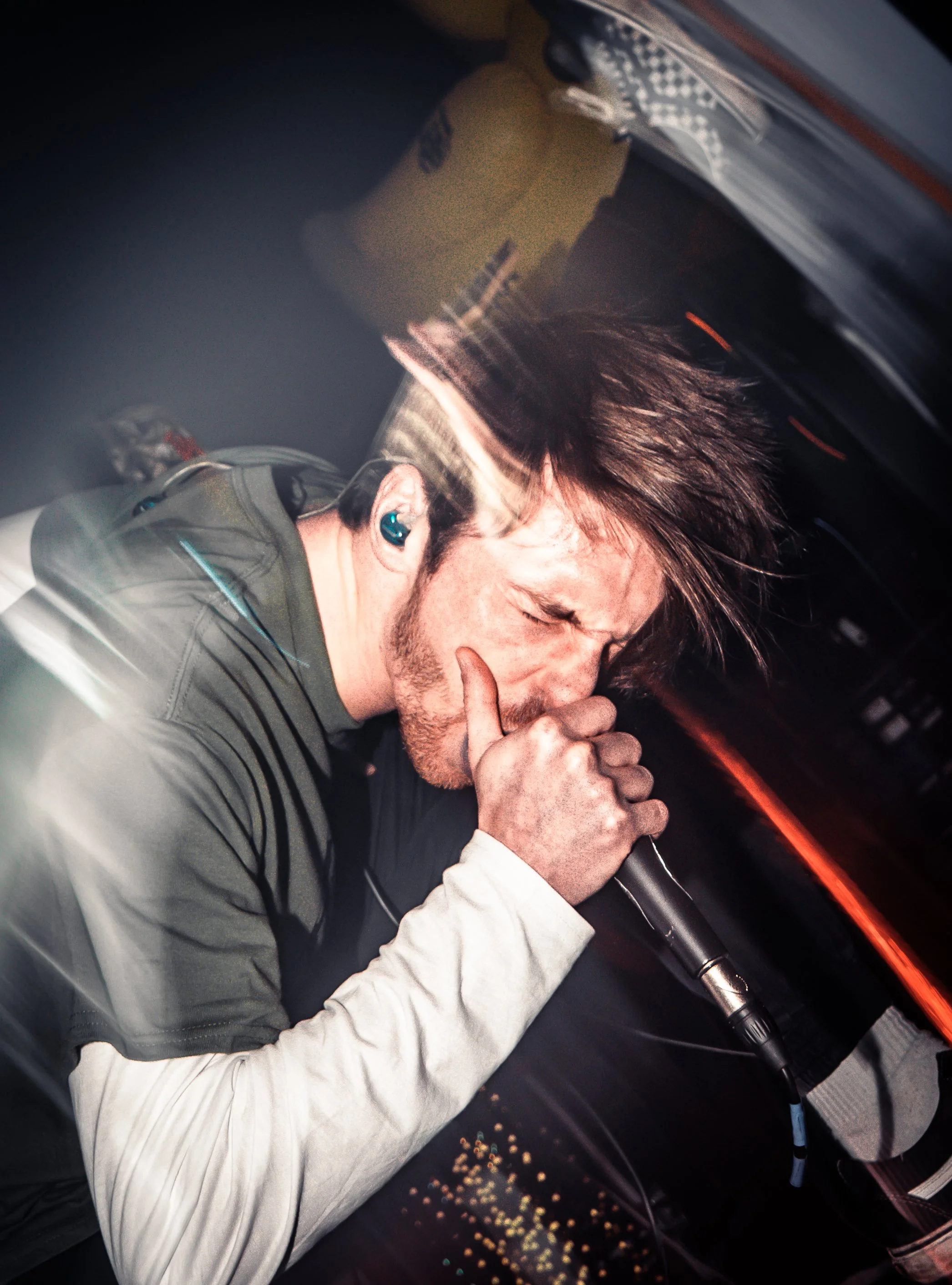 A man with brown hair and a beard singing into a microphone while wearing headphones inside a car at night.