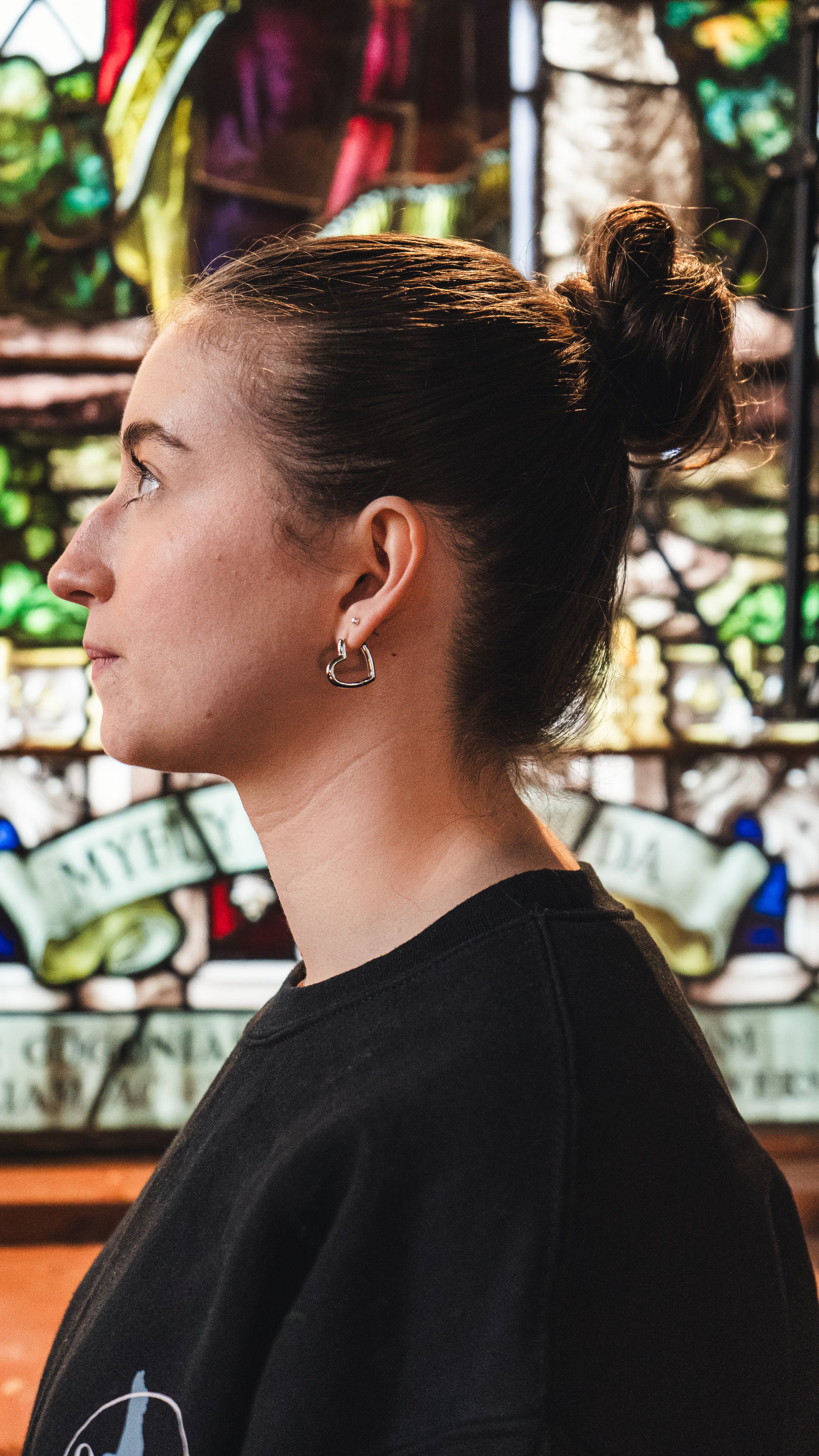 Profile of a young woman with dark brown hair tied up in a bun, wearing hoop earrings, standing in front of a colorful stained glass background.
