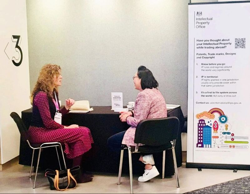 Two women sitting at a table having a discussion at a conference or meeting, with a sign related to intellectual property rights visible to their right.