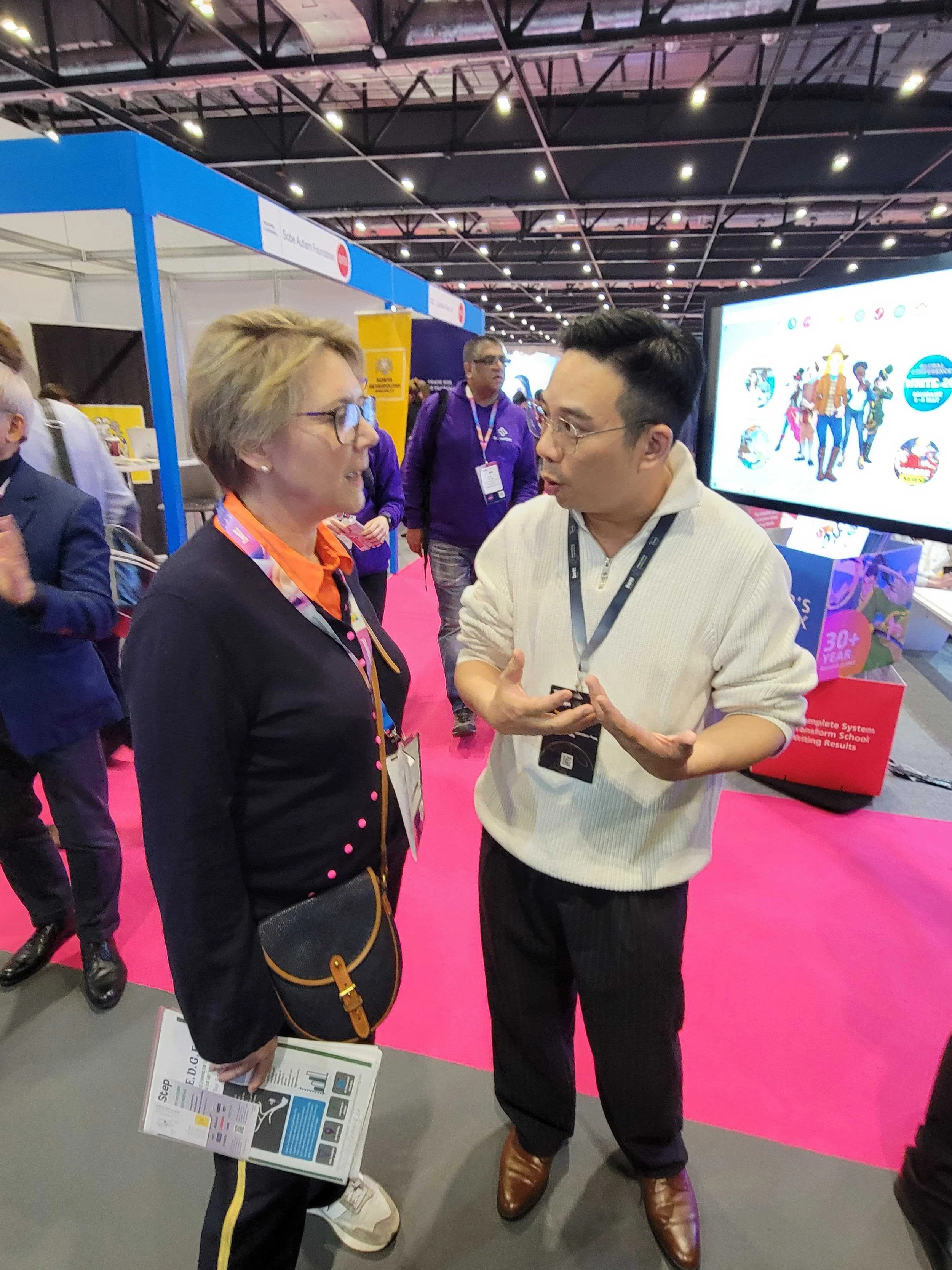 Two people in conversation at a trade show or conference, with a large screen in the background displaying cartoon characters and colorful graphics, and a bright pink carpeted floor.