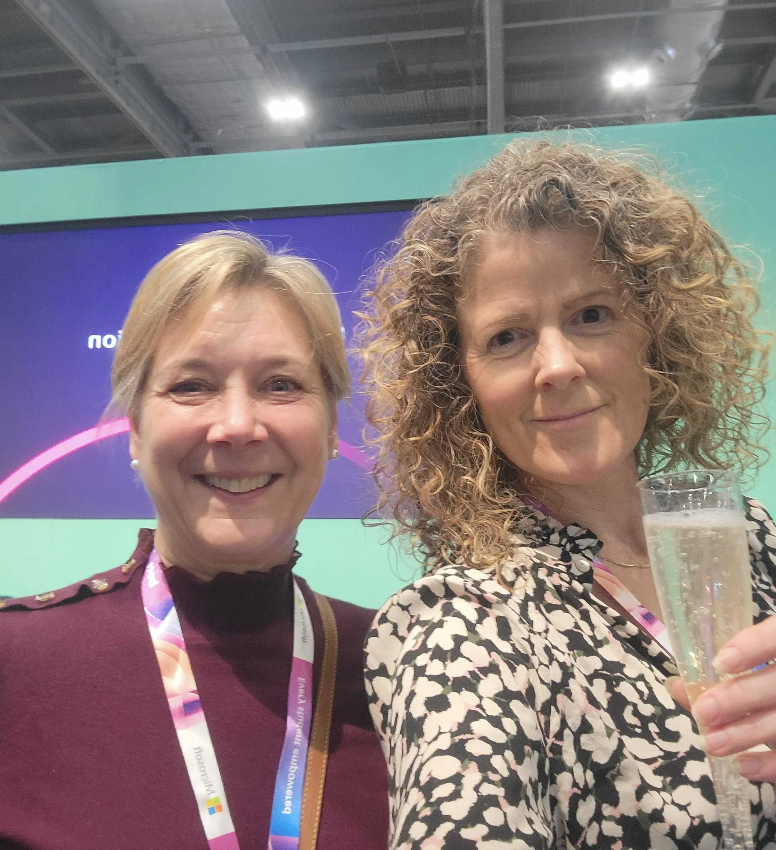 Two women taking a selfie at an indoor event, with one holding a glass of sparkling beverage, standing in front of a purple and teal backdrop.